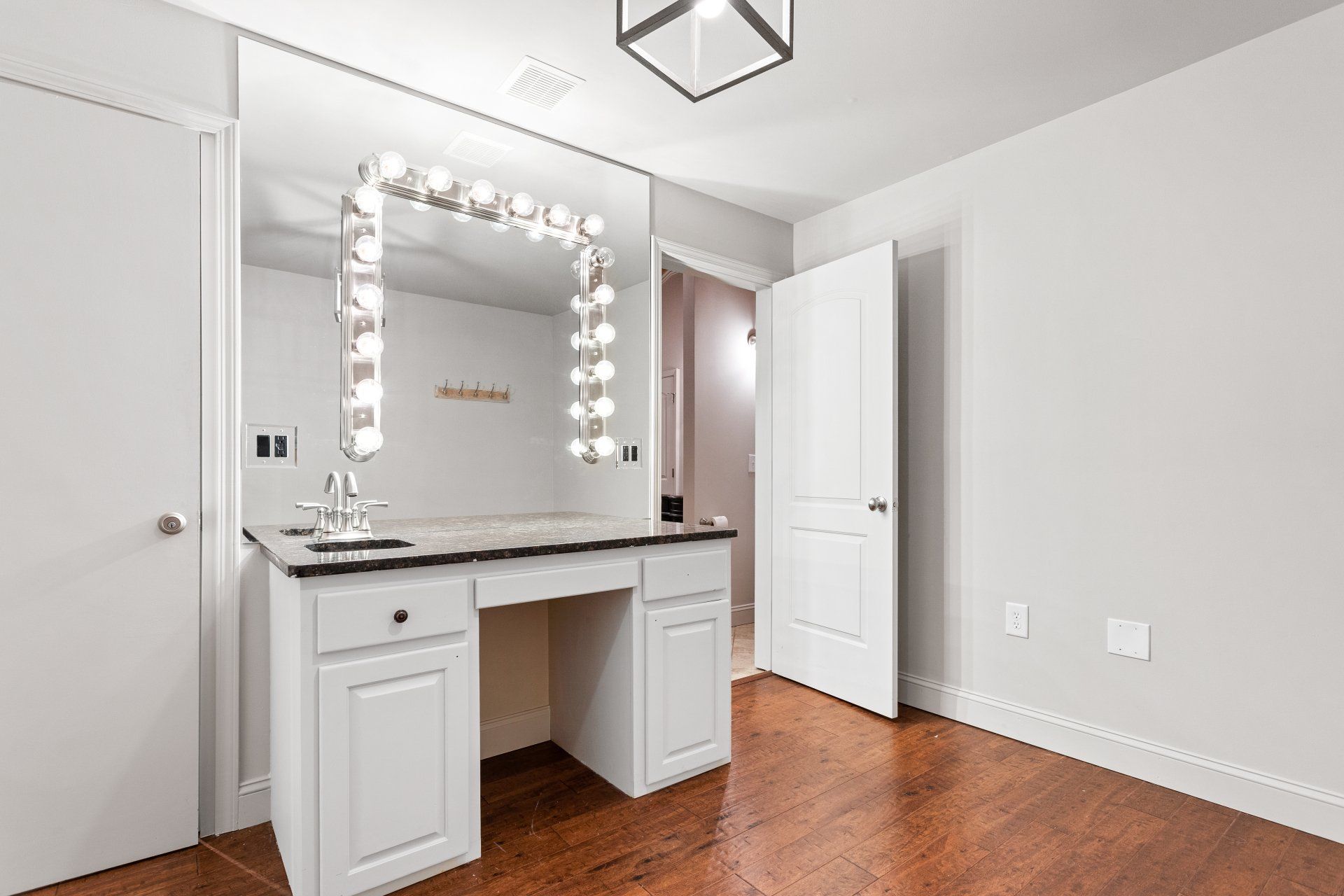Bright bedroom with white vanity, lighted mirror, hardwood floor, and open door