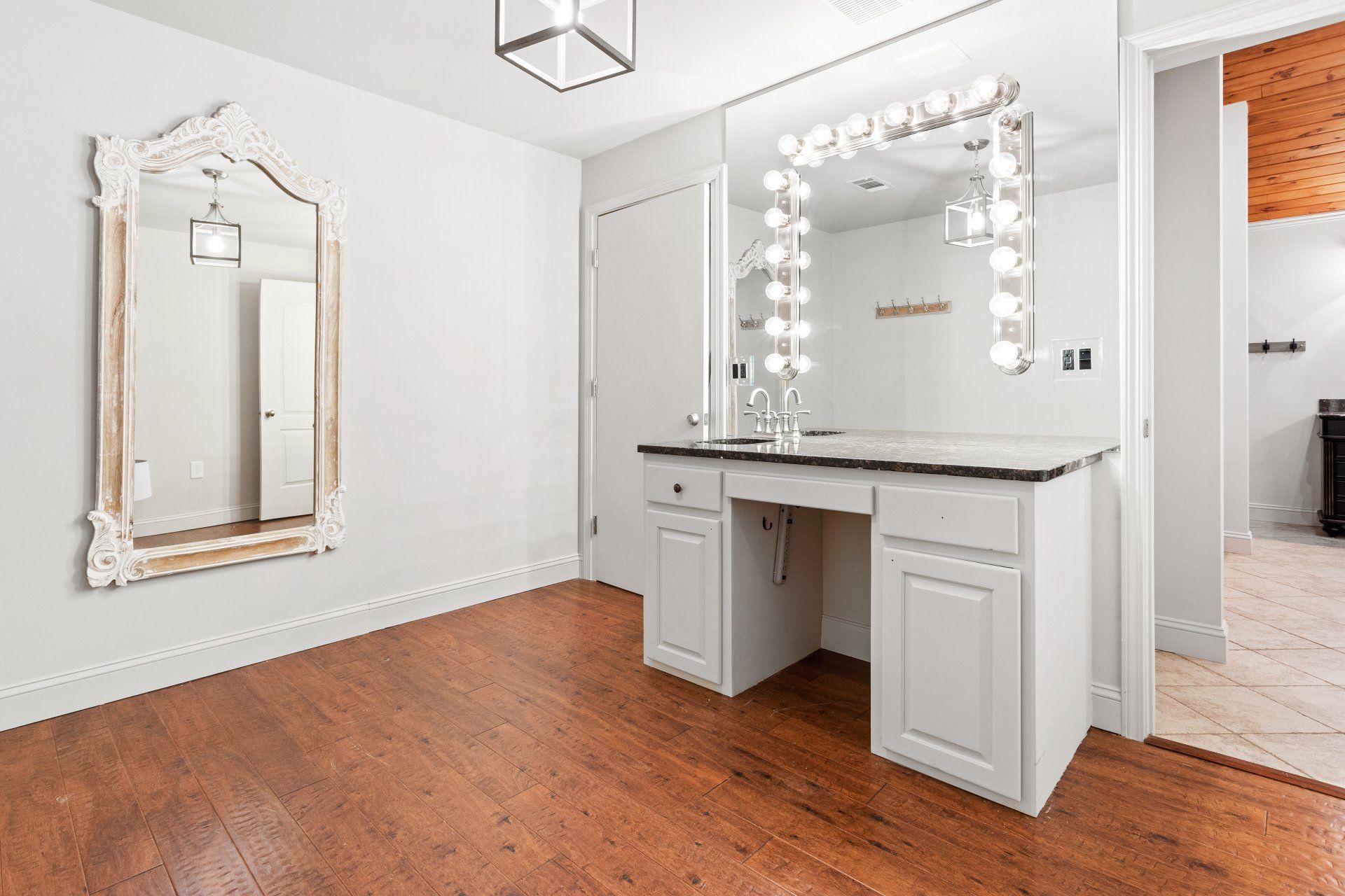 Bright vanity room with white cabinets, large lit mirror, ornate wall mirror, and wood floors
