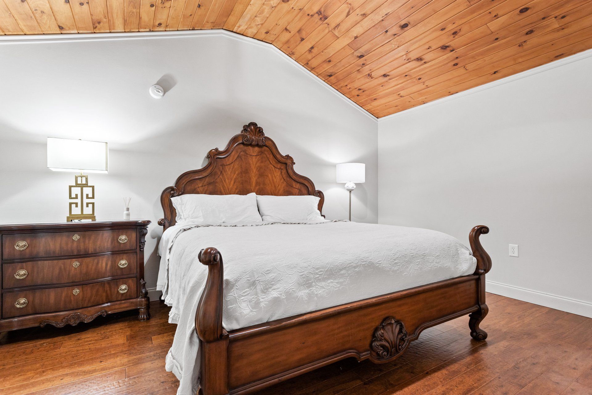 Elegant bedroom with ornate wooden bed, dresser, and angled wood-paneled ceiling