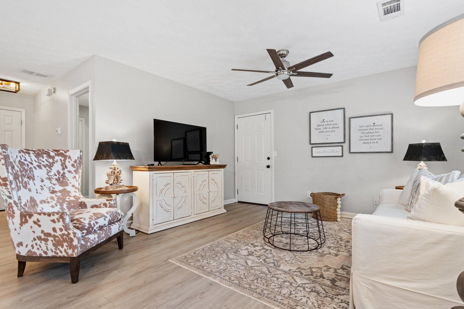 Bright living room with white sofa, patterned chair, TV console, coffee table, and ceiling fan