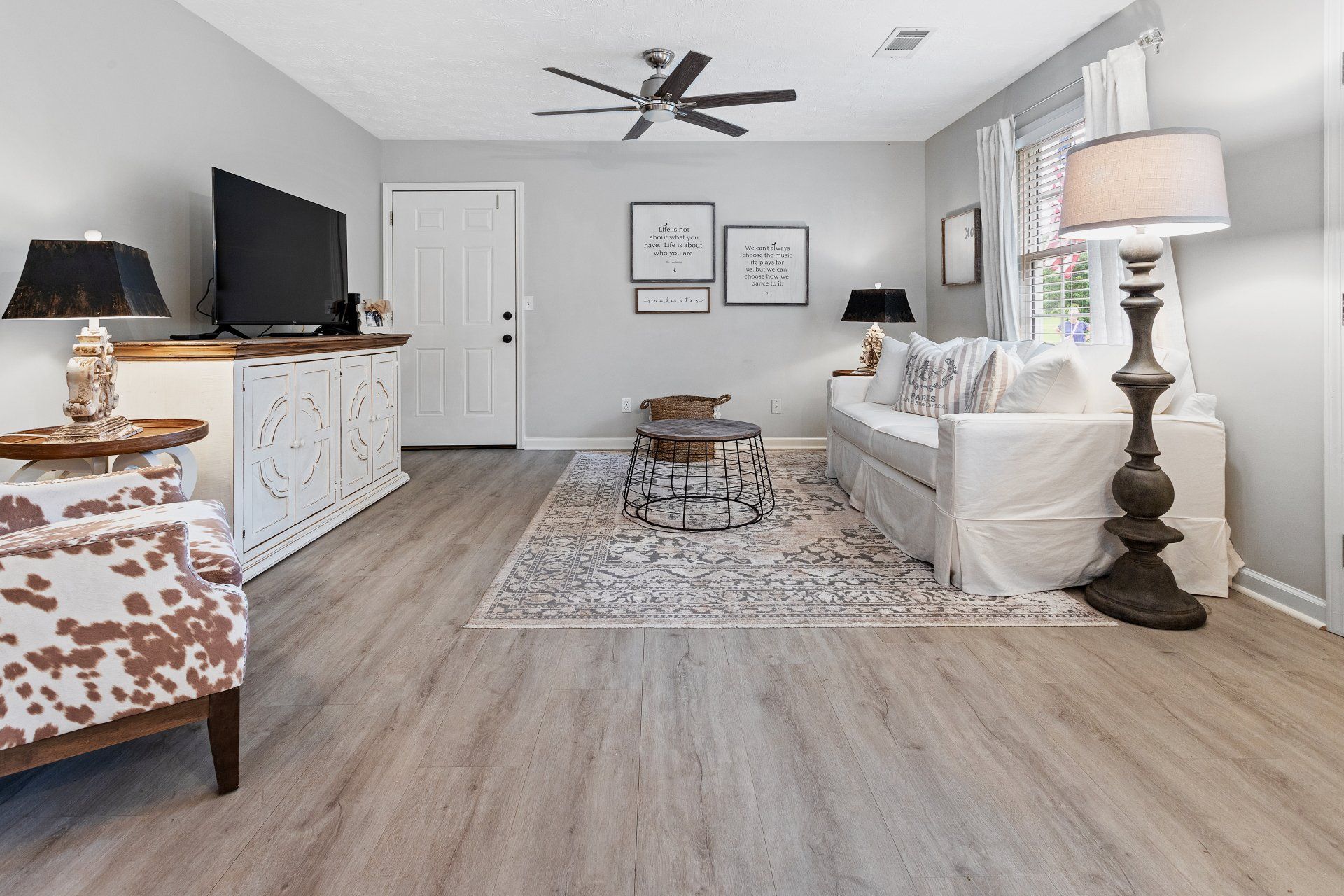 Bright living room with white sofa, patterned chair, TV console, ceiling fan, and large area rug