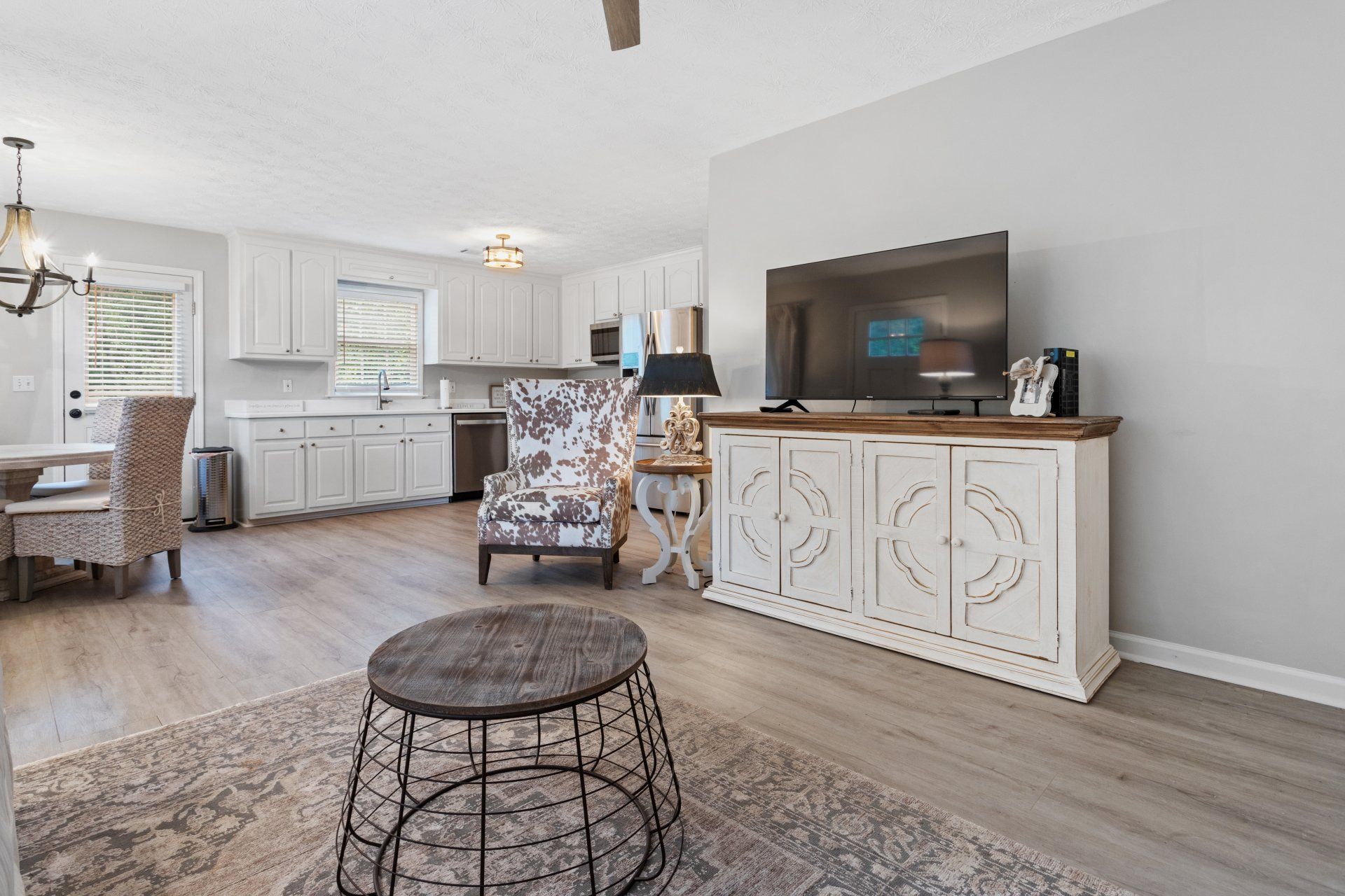 Bright open living room with white kitchen, patterned chair, TV console, and wire coffee table on a rug