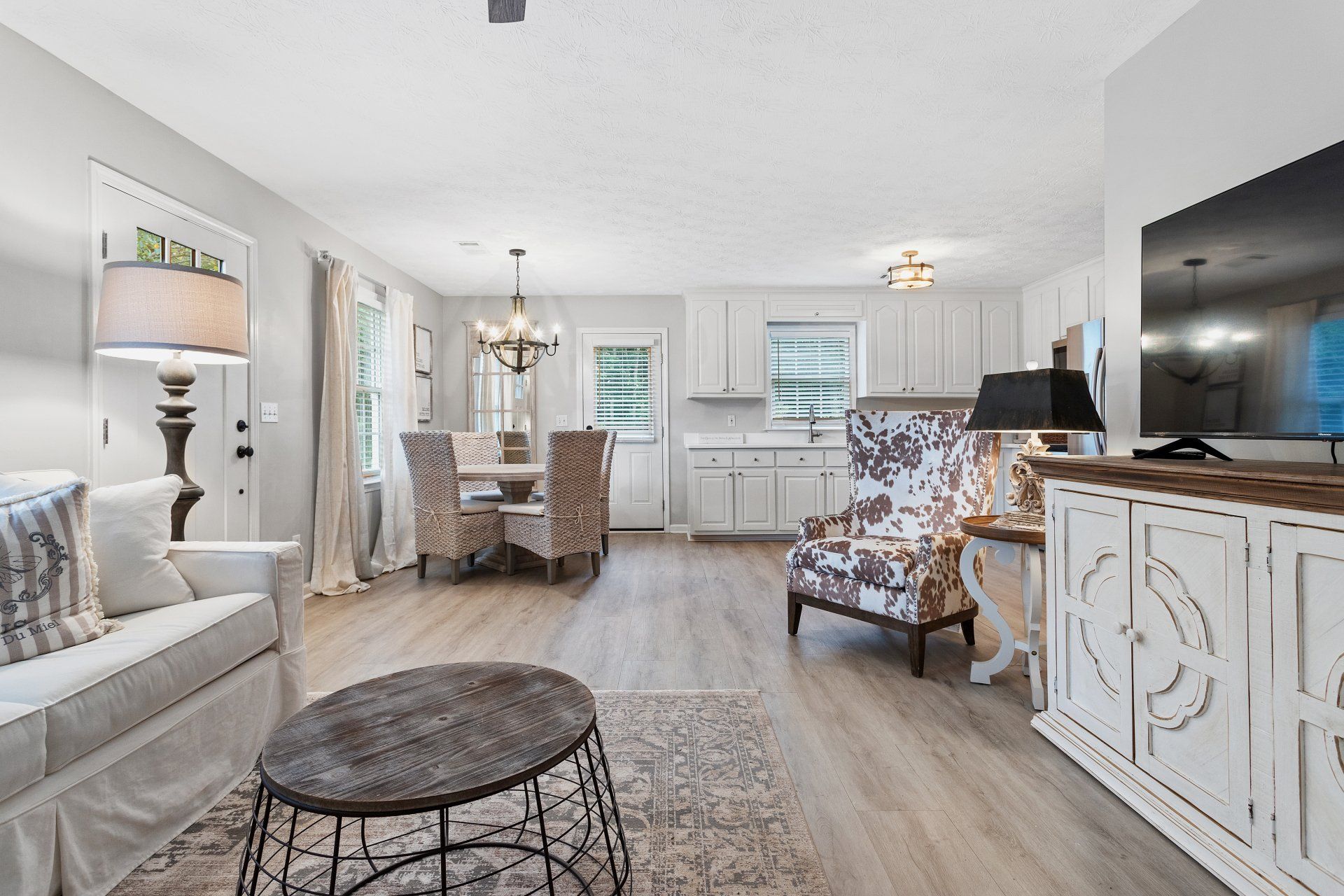 Bright open-concept living room and dining area with white furniture, wood floors, and large windows.