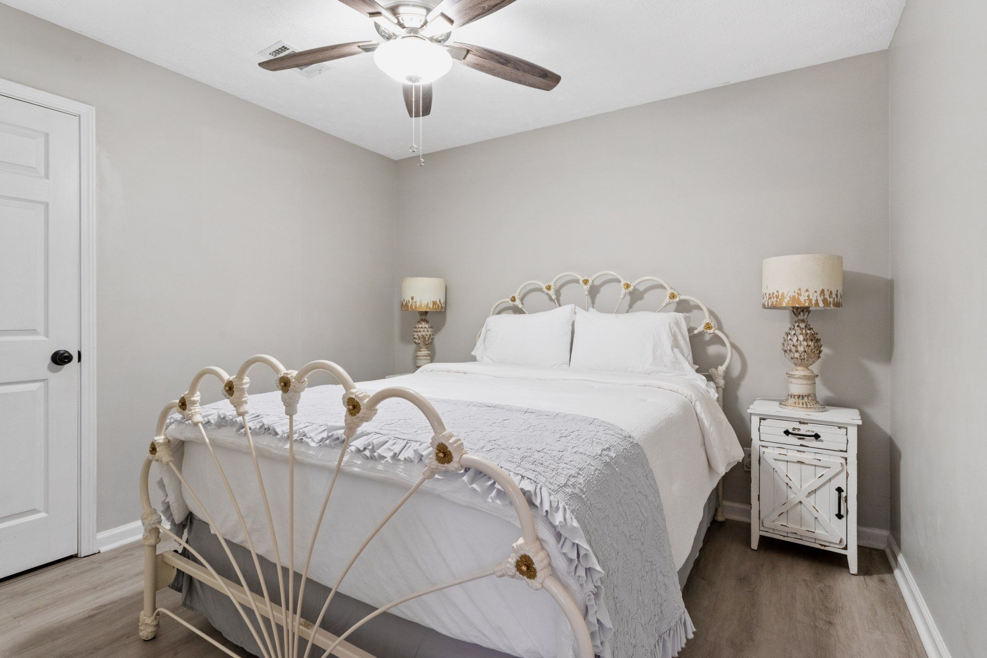 Bright bedroom with white bedding, gold-accented bedframe, nightstands, and a ceiling fan