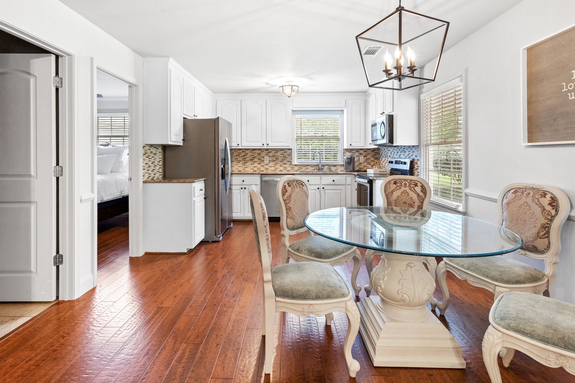 Bright dining and kitchen area with glass table, wicker chairs, hardwood floors, and white cabinets