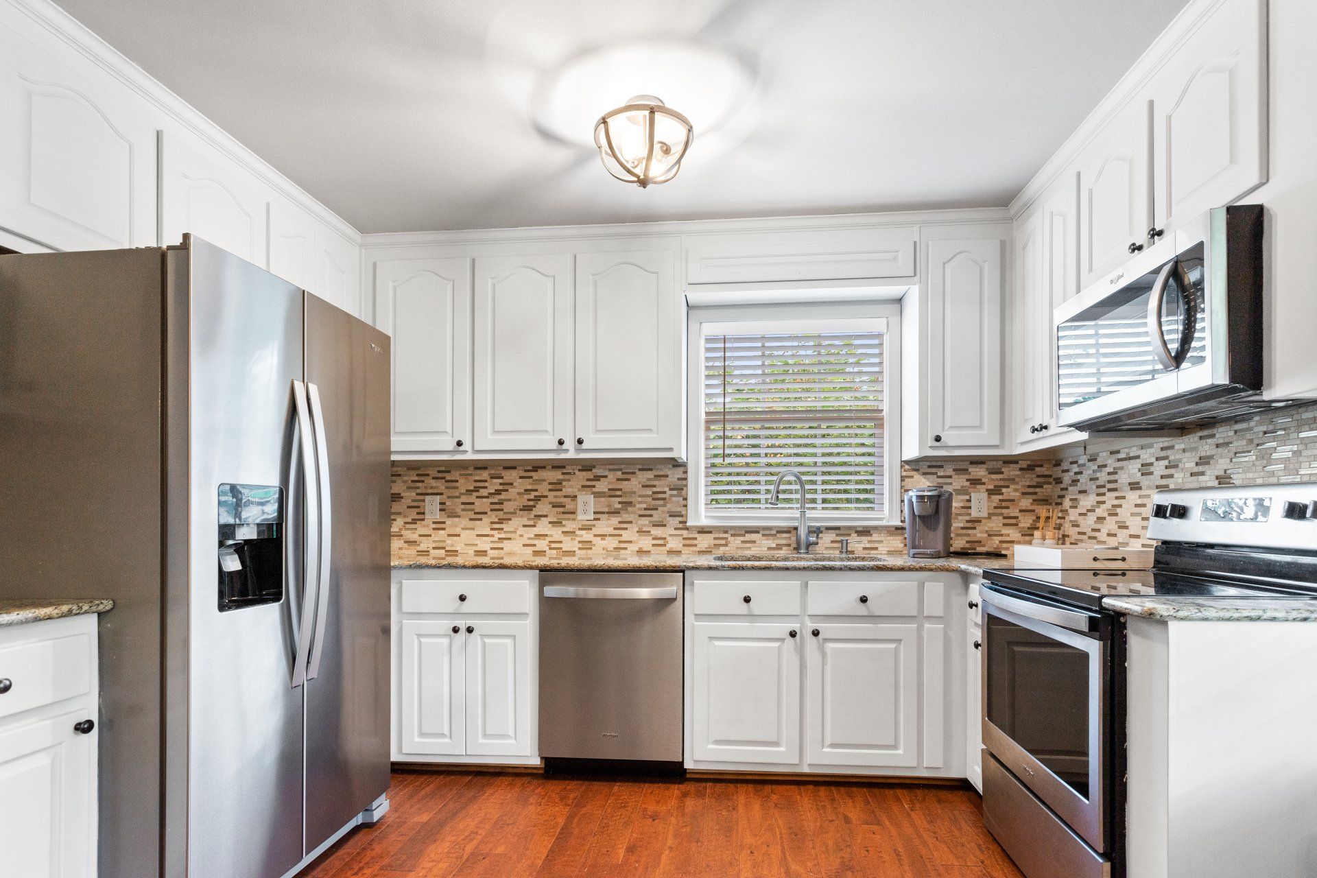 Bright white kitchen with stainless steel appliances, wood floor, and tiled backsplash around a window