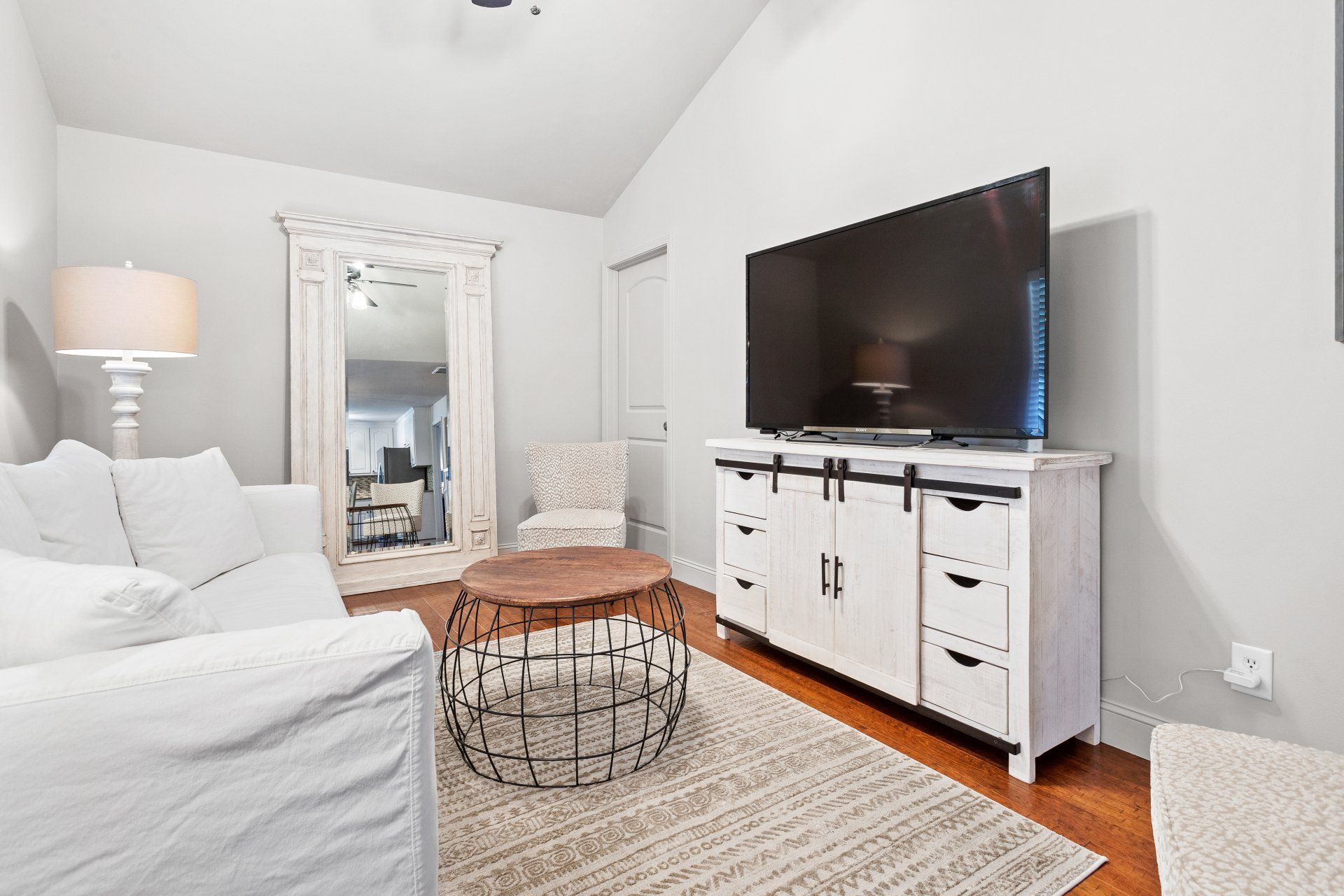 Bright living room with white sofa, wall-mounted TV, mirror, lamp, and round coffee table on a rug.