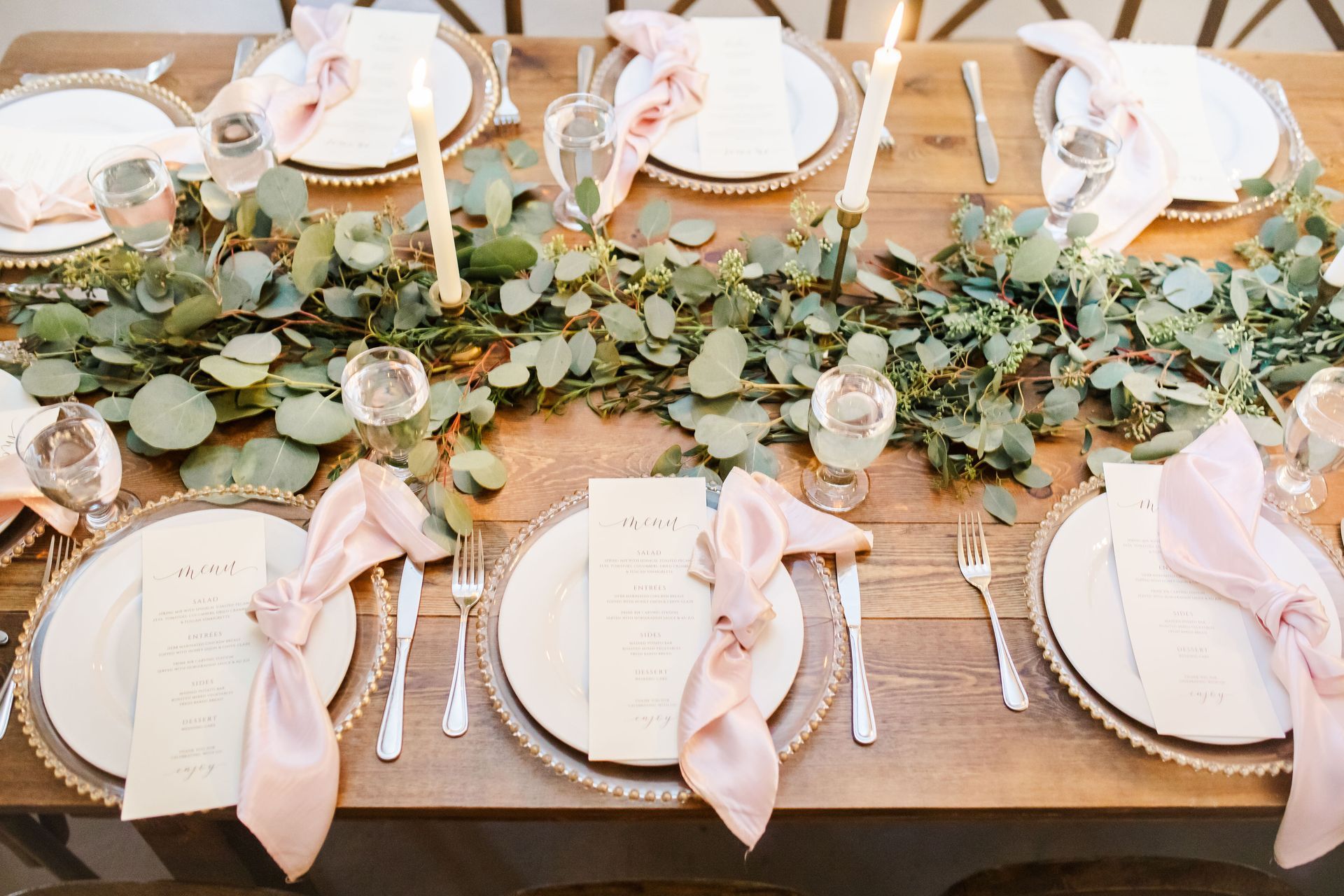 Elegant table setting with white plates, pink napkins, greenery garland, and glassware on a wooden table
