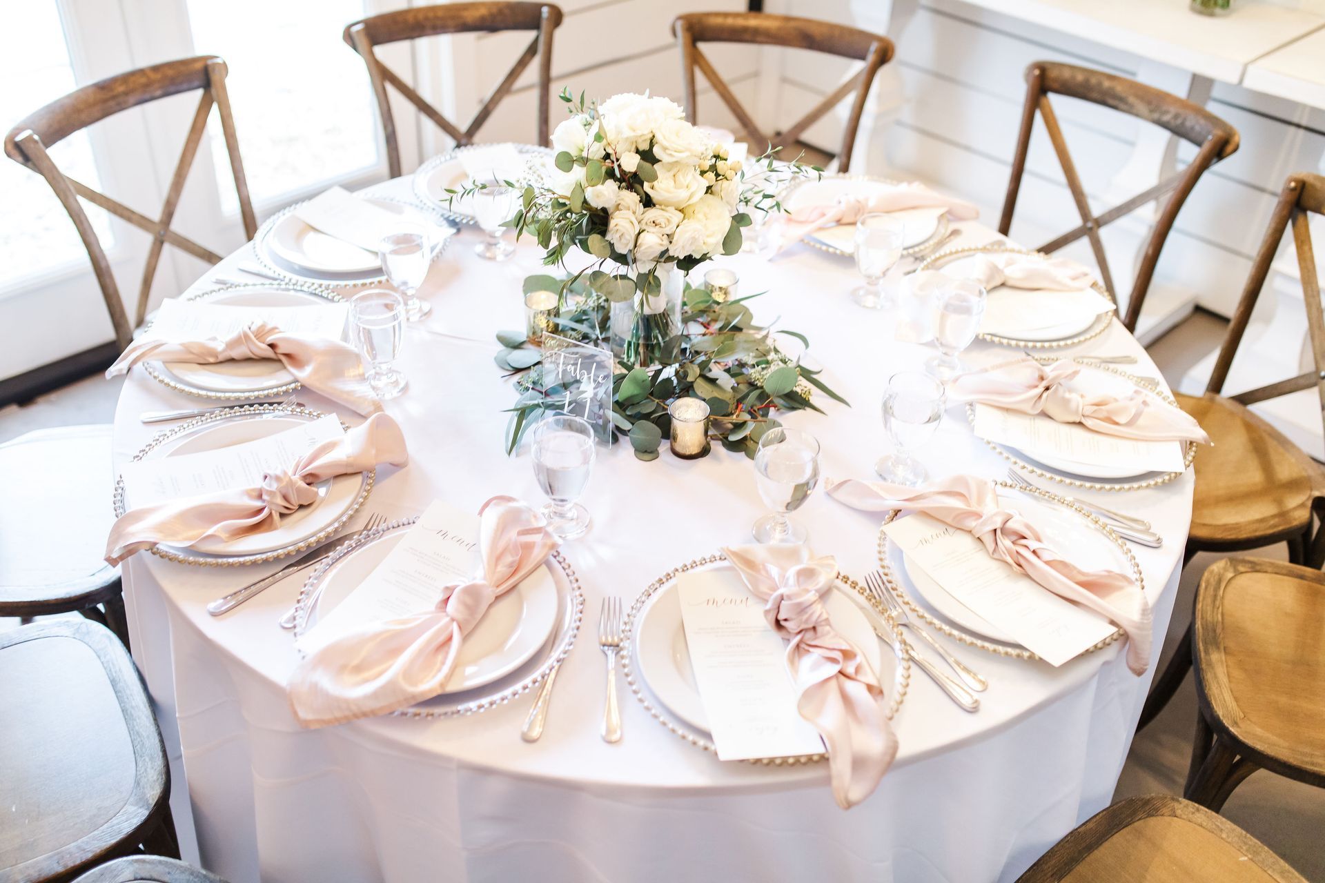 Round white table set for a wedding reception with blush napkins, gold accents, and floral centerpiece