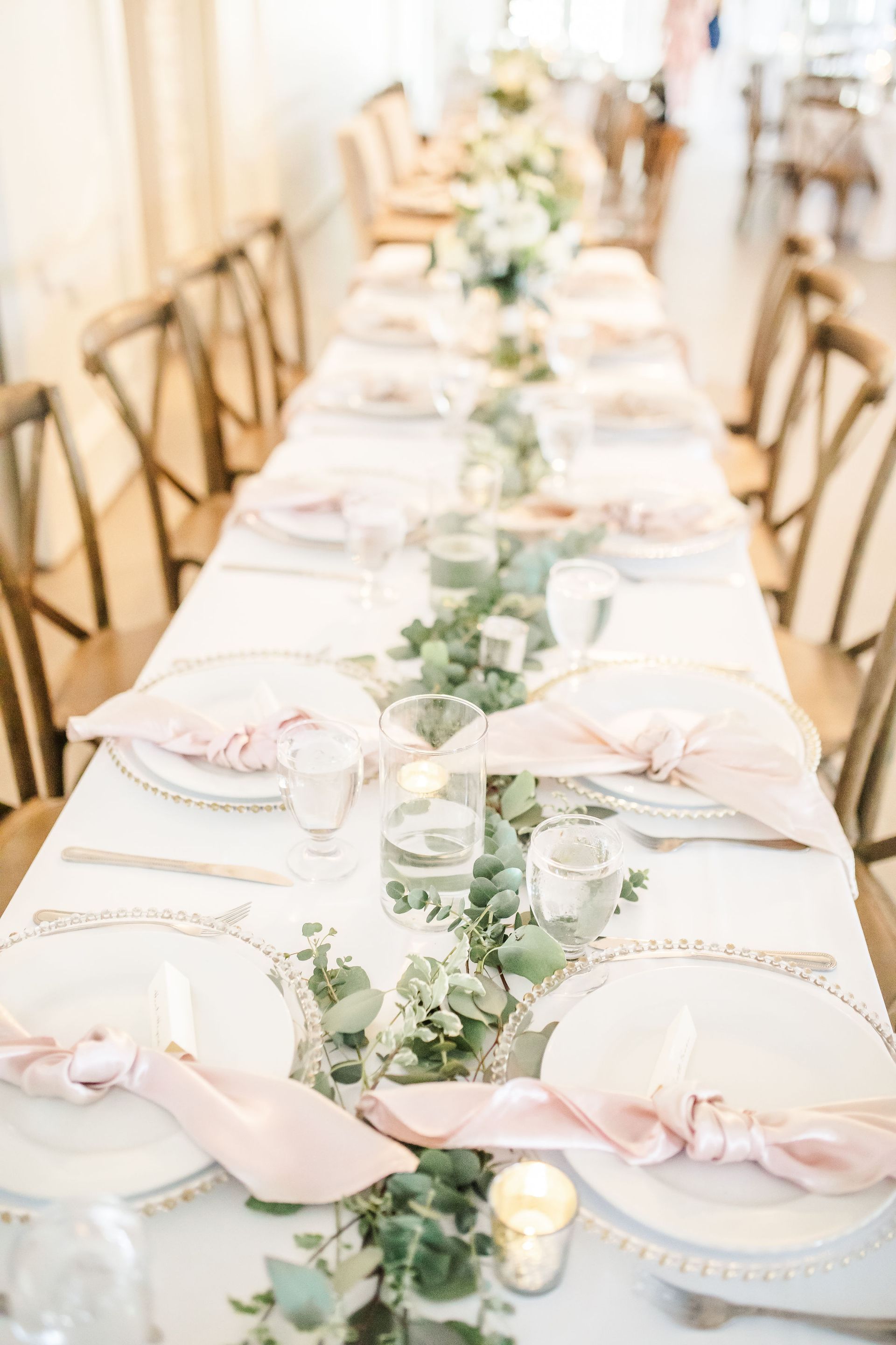 Long banquet table set with white linens, blush napkins, candles, and greenery centerpieces