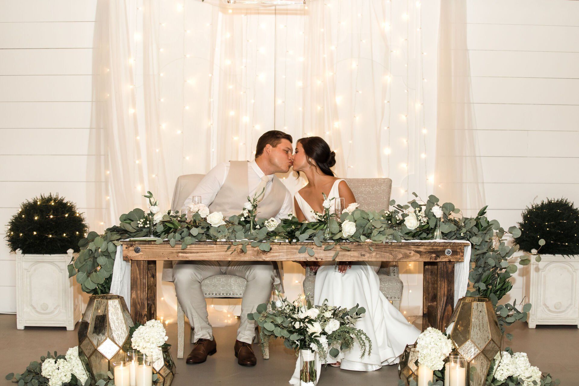 Couple kissing at a rustic wedding table with white flowers and soft fairy lights