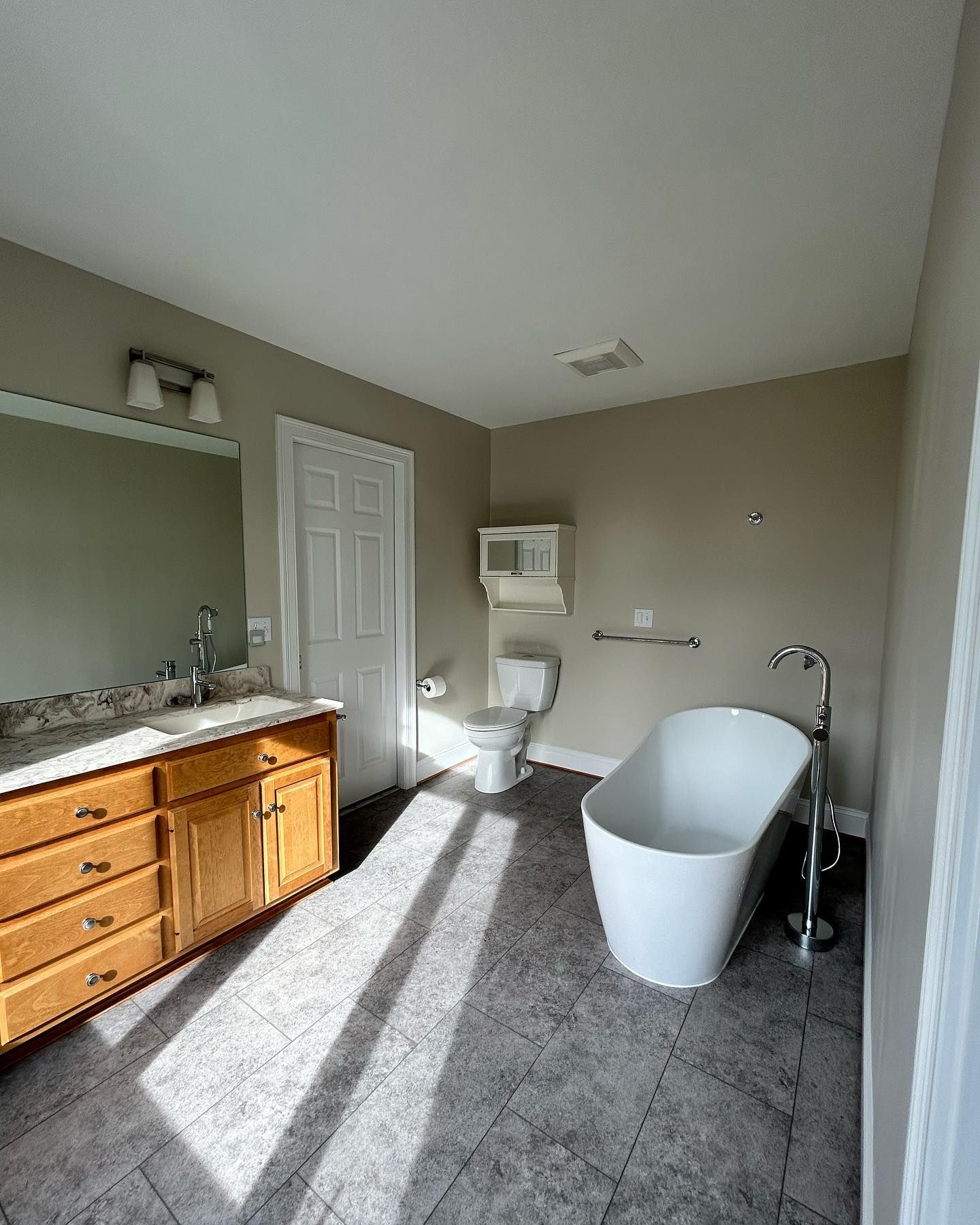Bathroom with freestanding tub, vanity, and toilet. Gray tile floor, neutral walls. Sunlight streams in.