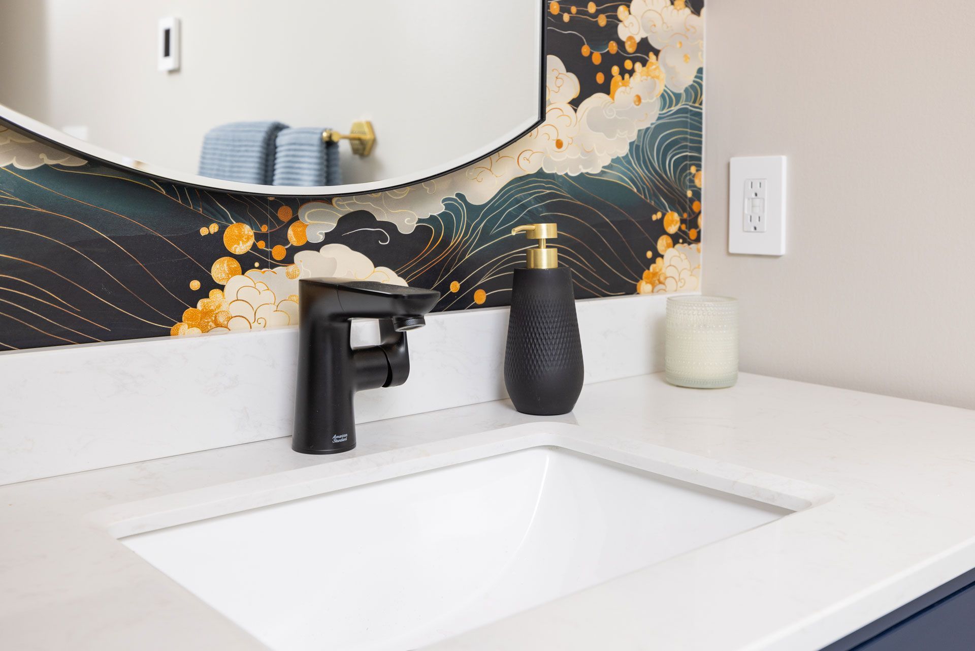 Bathroom sink with black faucet, soap dispenser, and a candle on a white countertop. Floral wallpaper and round mirror.
