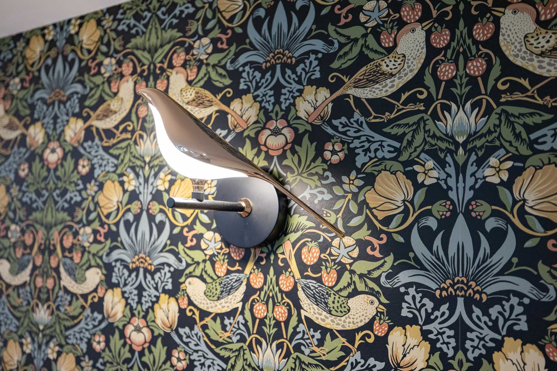 Bird-shaped wall sconce with warm light, mounted on patterned wallpaper featuring birds and floral elements.