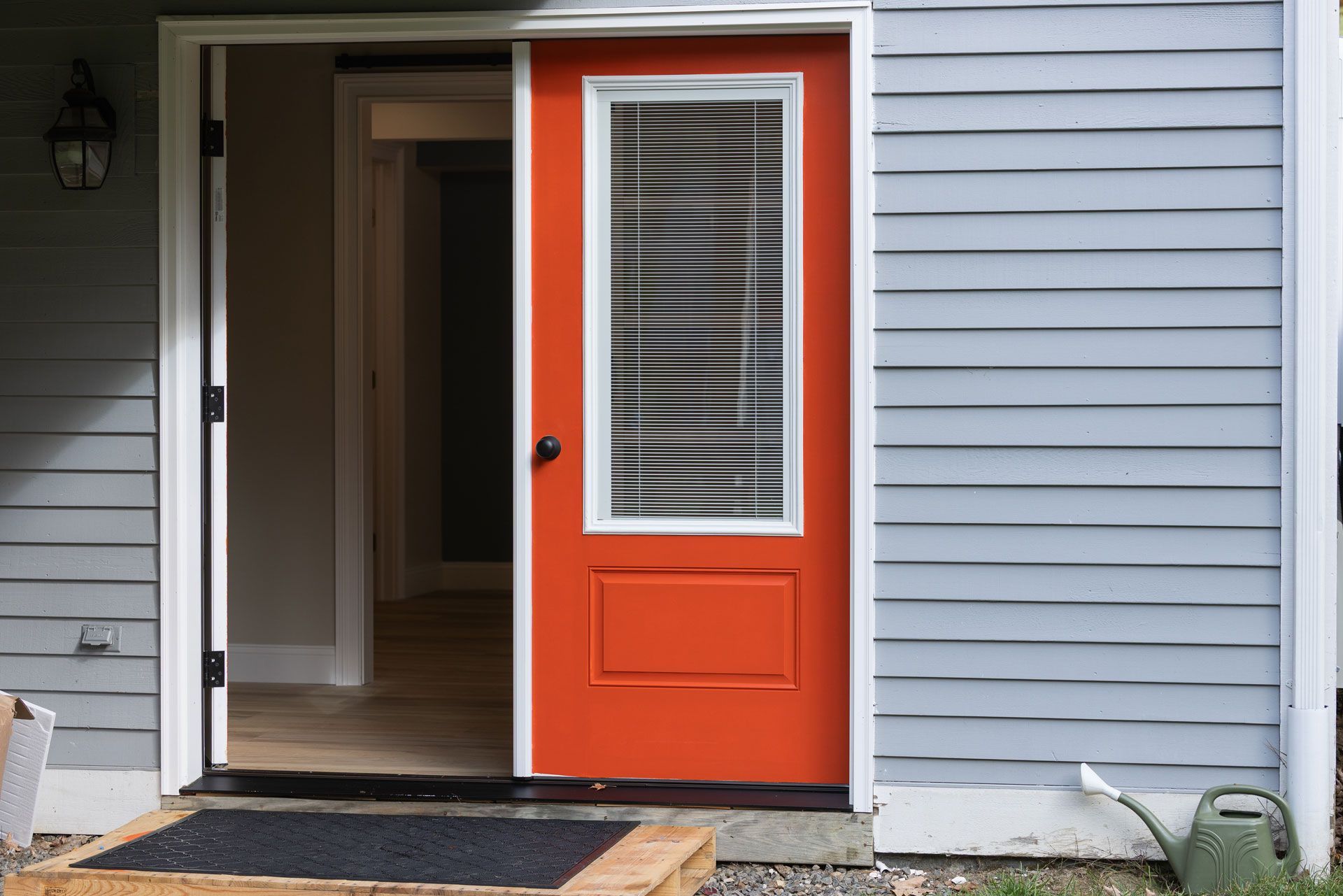 Orange door with window and blinds, open on a gray house, doorway to another room.