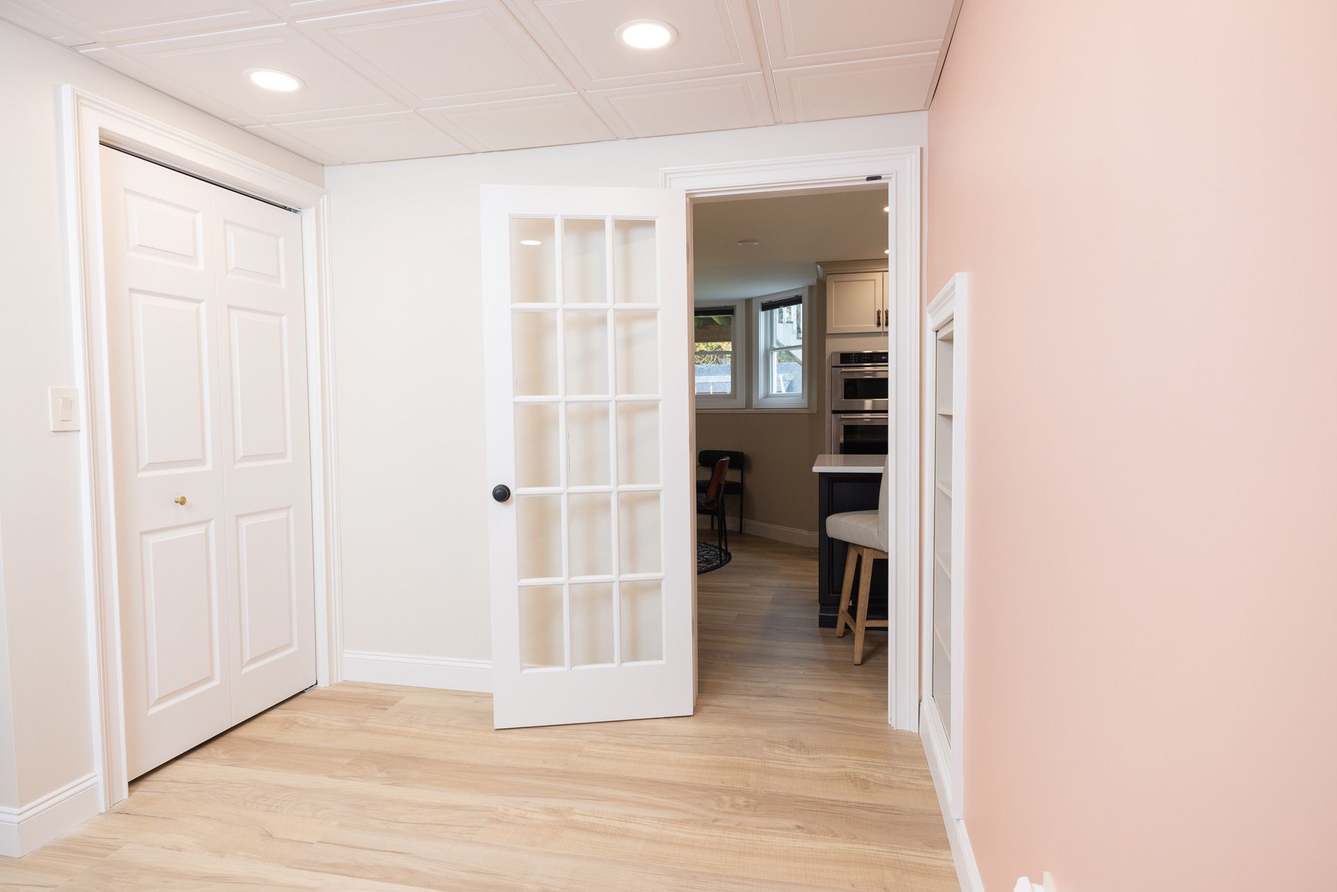A white French door opens to a kitchen, beside a white closet and a pink wall in a room with light wood floors.