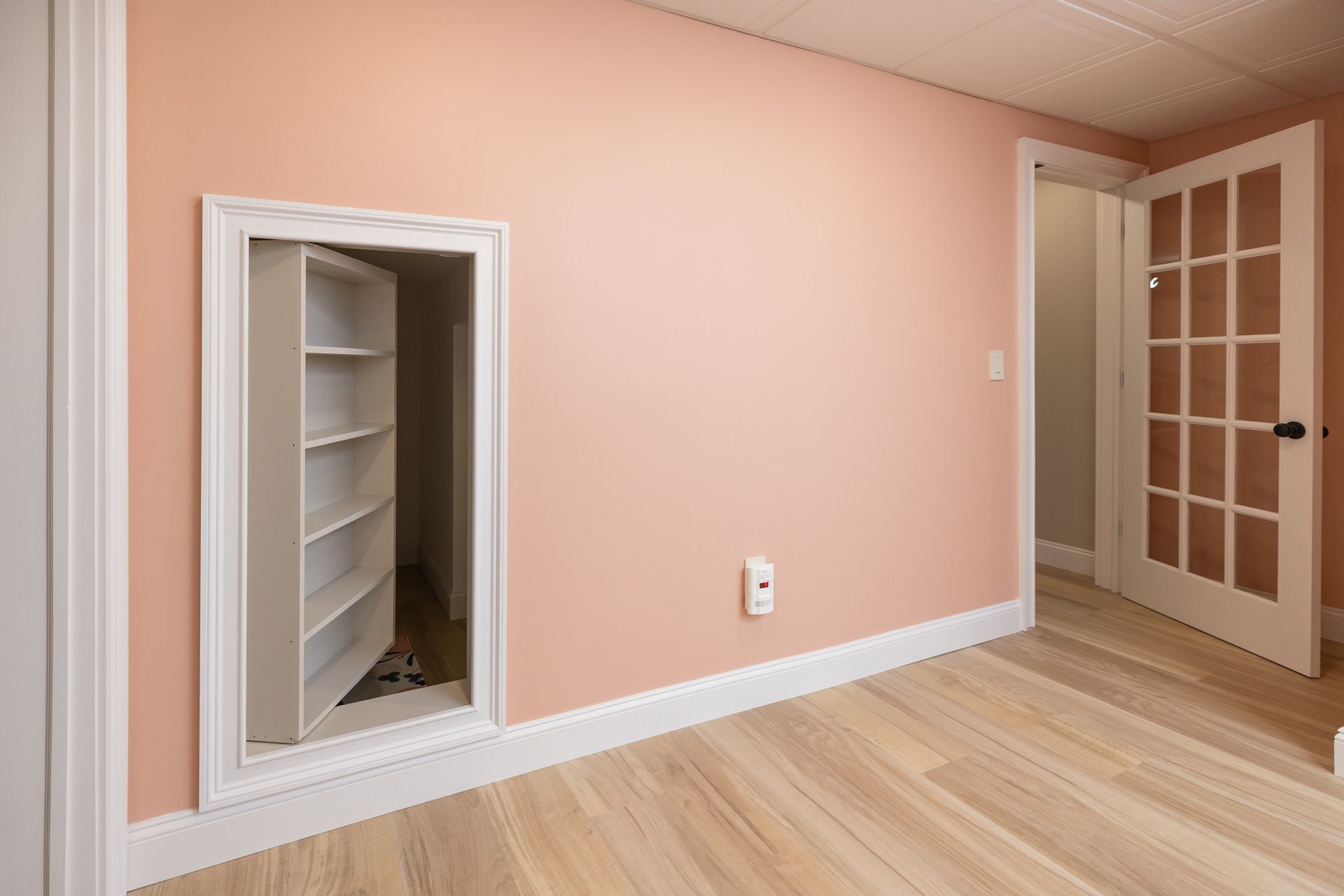 A pink-walled room with a hidden shelving unit behind a door; light wood floors, a door, and white trim.