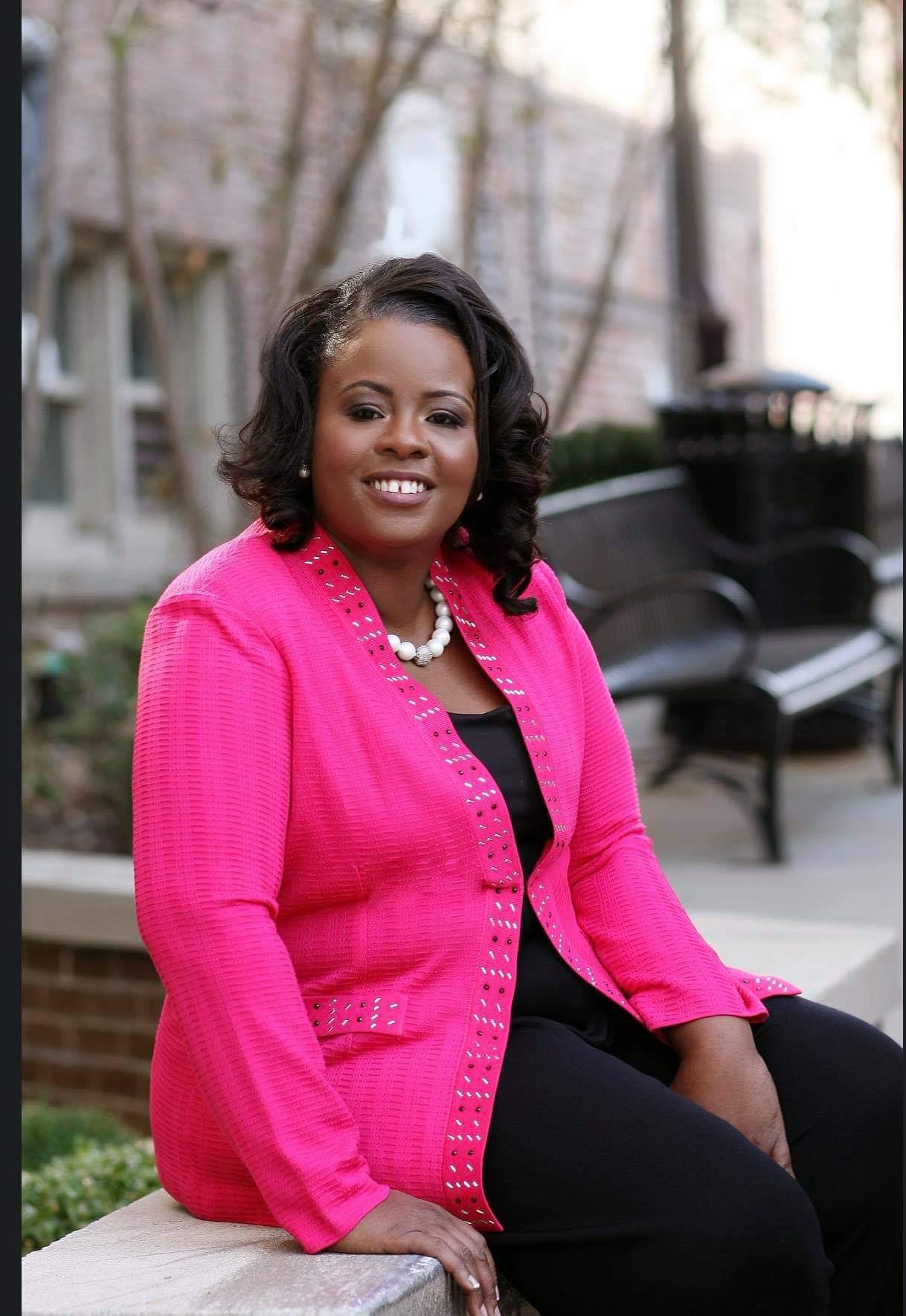 A woman in a pink jacket and black pants is sitting on a wall.