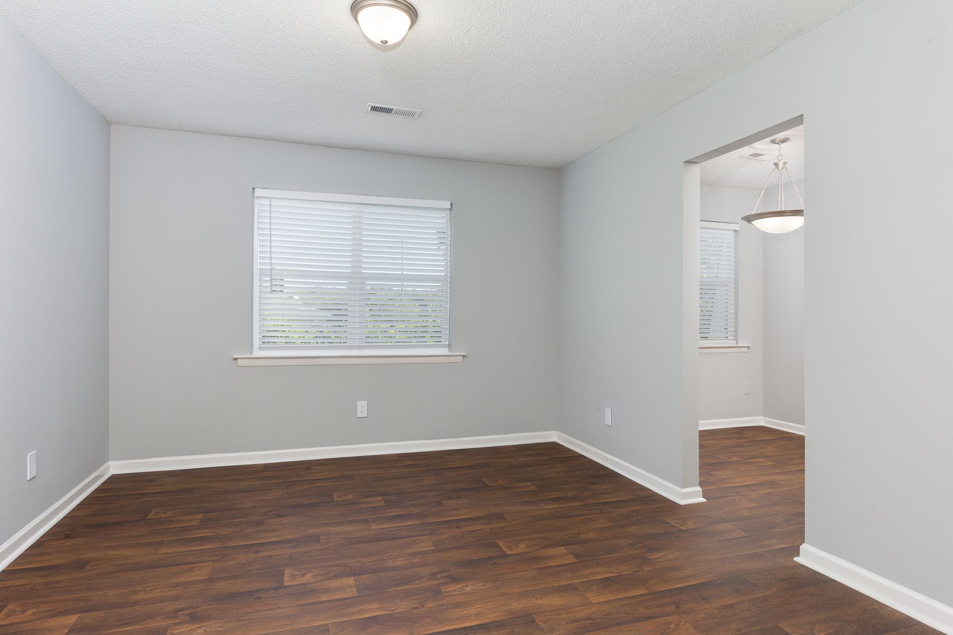 Empty room with light gray walls, dark wood-look floors, a single window with blinds, and a doorway to a lit hallway.