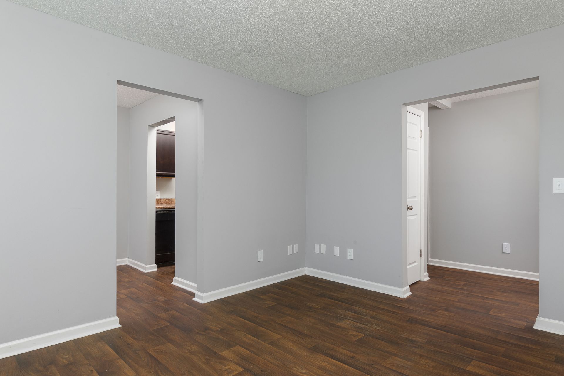 An empty room with light gray walls, white trim, wood-look flooring, and doorways leading to a kitchen and a hallway.