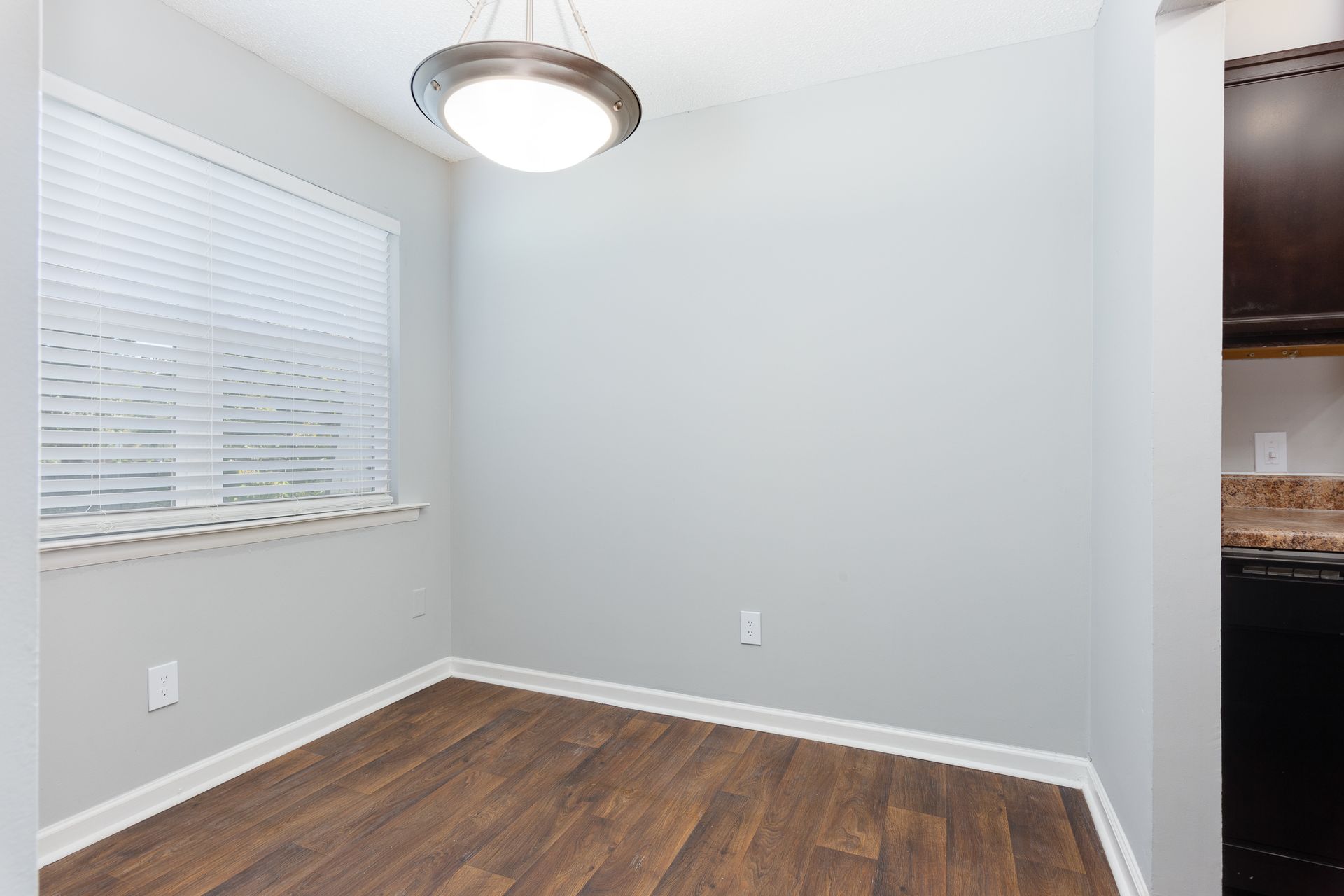A small dining area with light gray walls, dark wood-look floors, a large window with white blinds, and a pendant light.