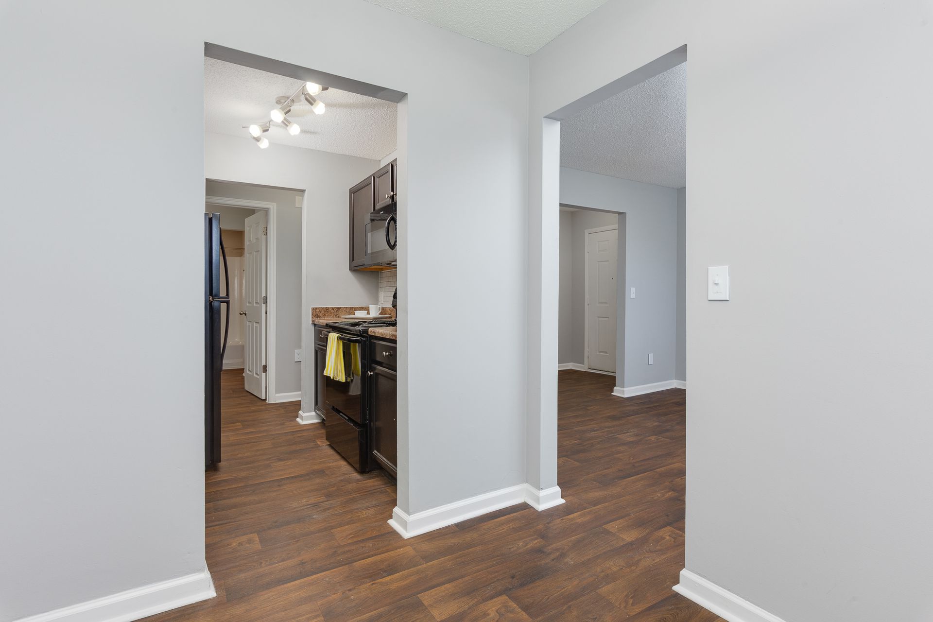 Interior view of a hallway featuring gray walls, wood-look flooring, and doorways opening into a kitchen and another room.