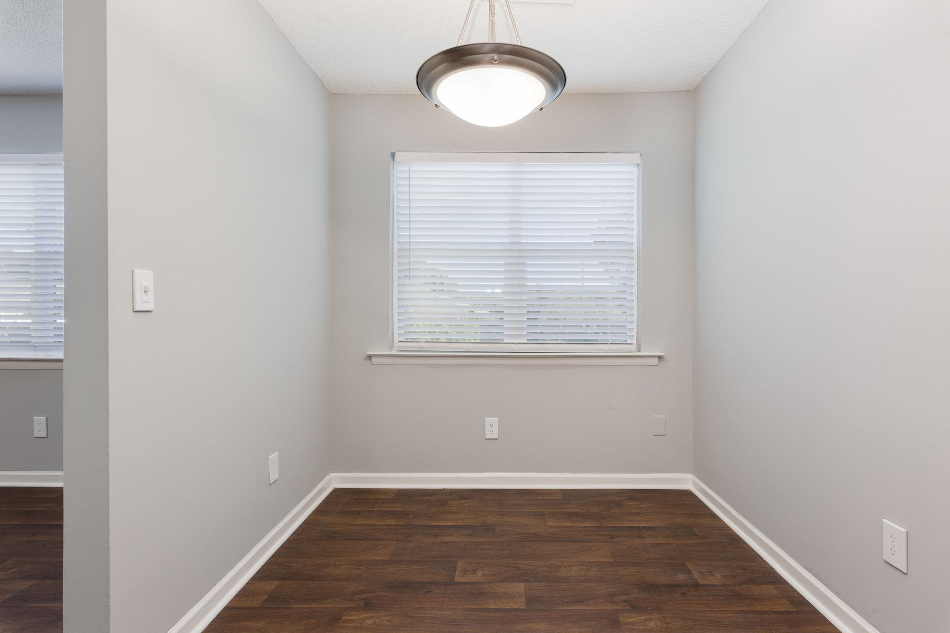 Empty dining area with light gray walls, dark wood-look floors, a white window with blinds, and a central pendant light.