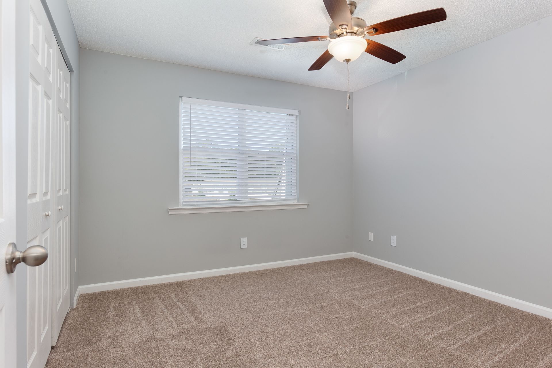 An empty room with light gray walls, brown carpet, a white door, a window with closed blinds, and a ceiling fan.