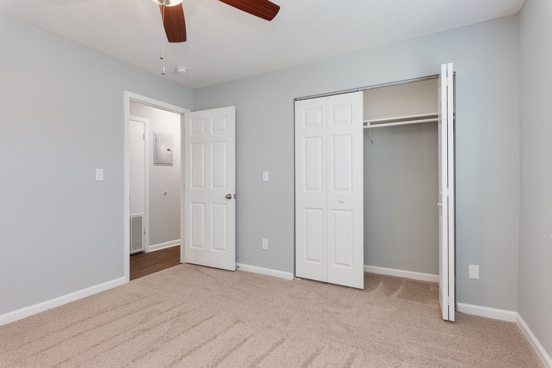 An empty room with light gray walls, tan carpet, a ceiling fan, a standard bedroom door, and an open closet.