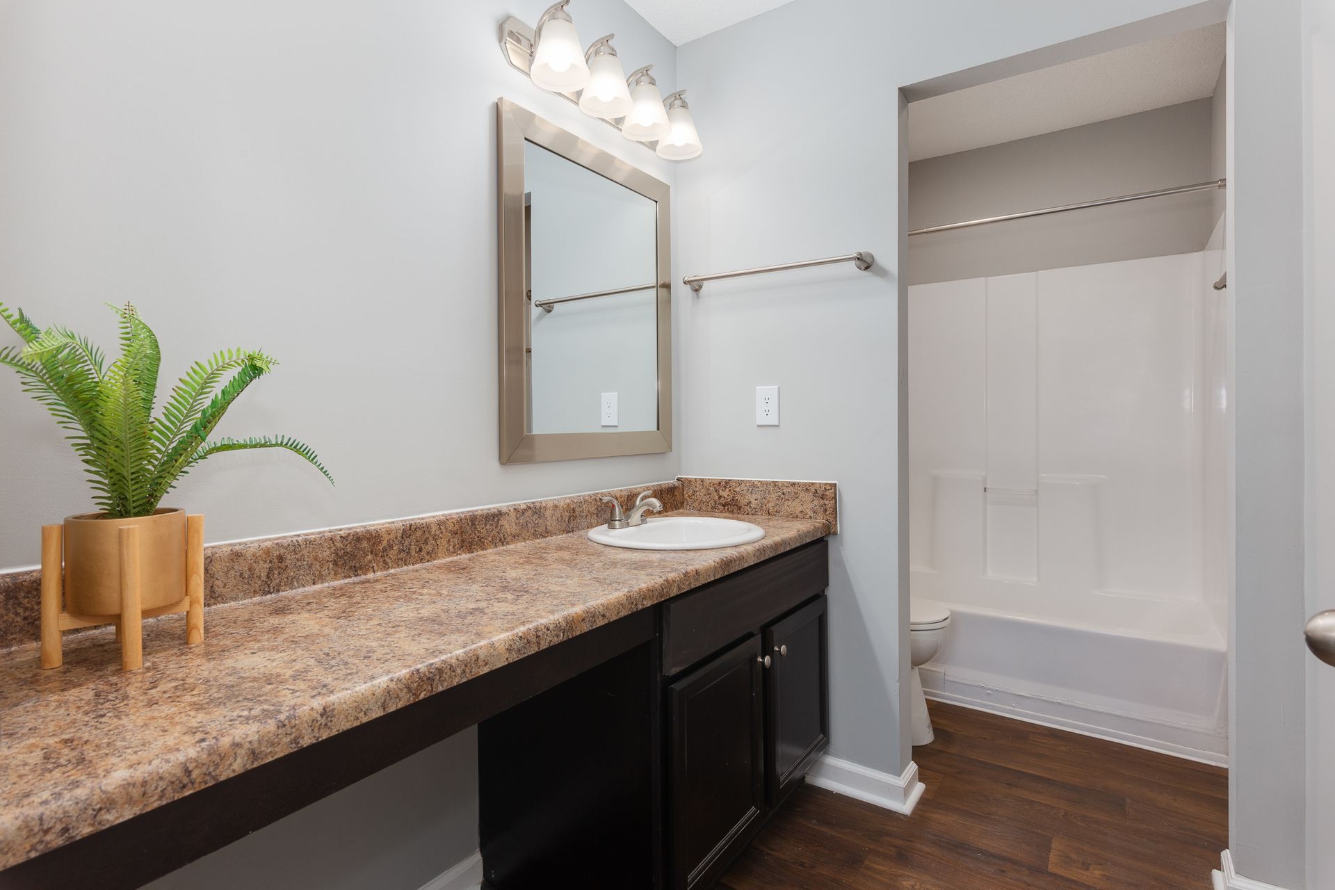 A bathroom with dark brown cabinets, tan speckled countertop, wall mirror, vanity lighting, and a white shower tub.