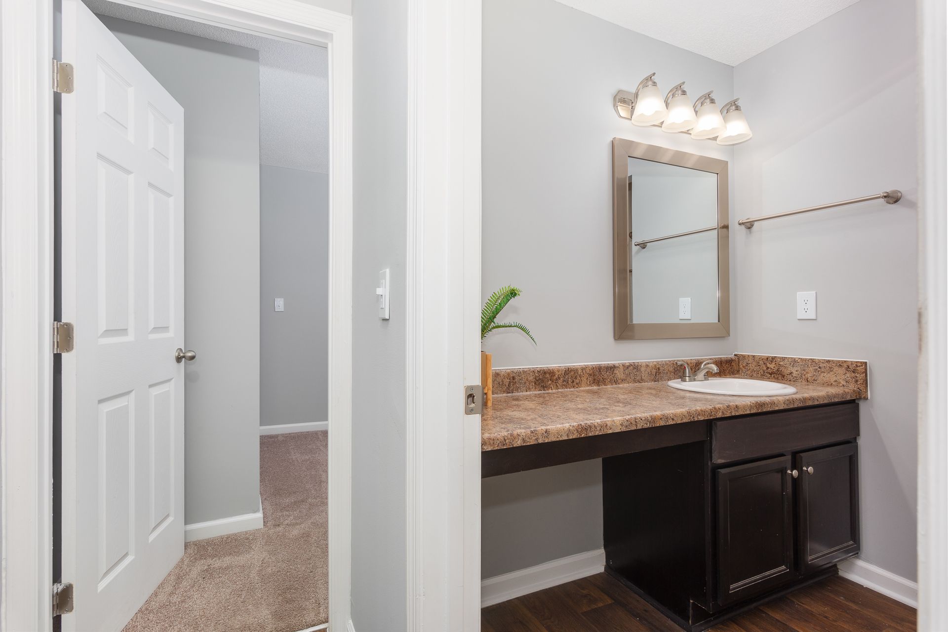 A bathroom vanity with dark wood cabinets, a speckled countertop, and mirror next to a doorway leading to a carpeted room.