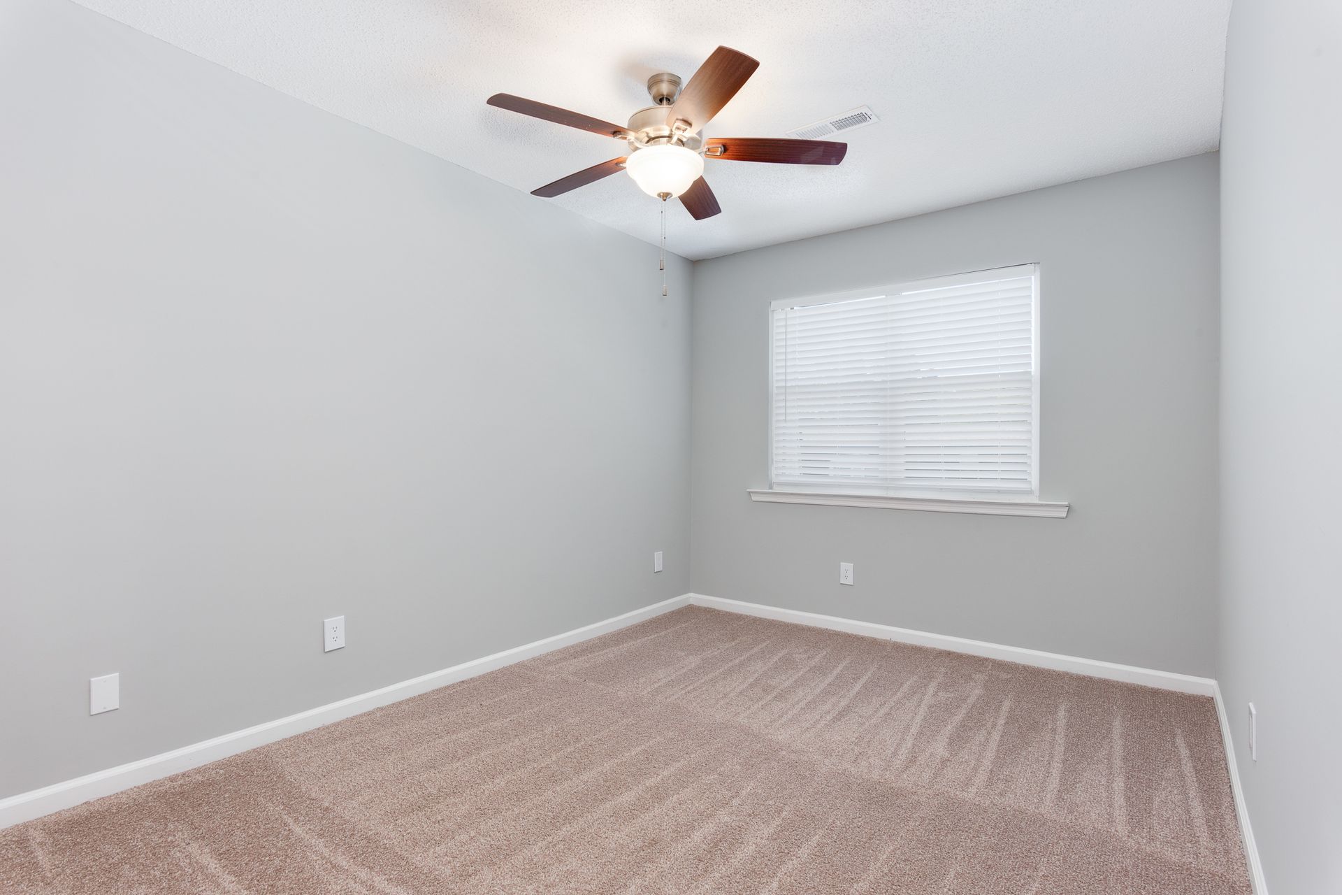 A bright, empty bedroom with light gray walls, carpeted flooring, a ceiling fan, and a single window with white blinds.