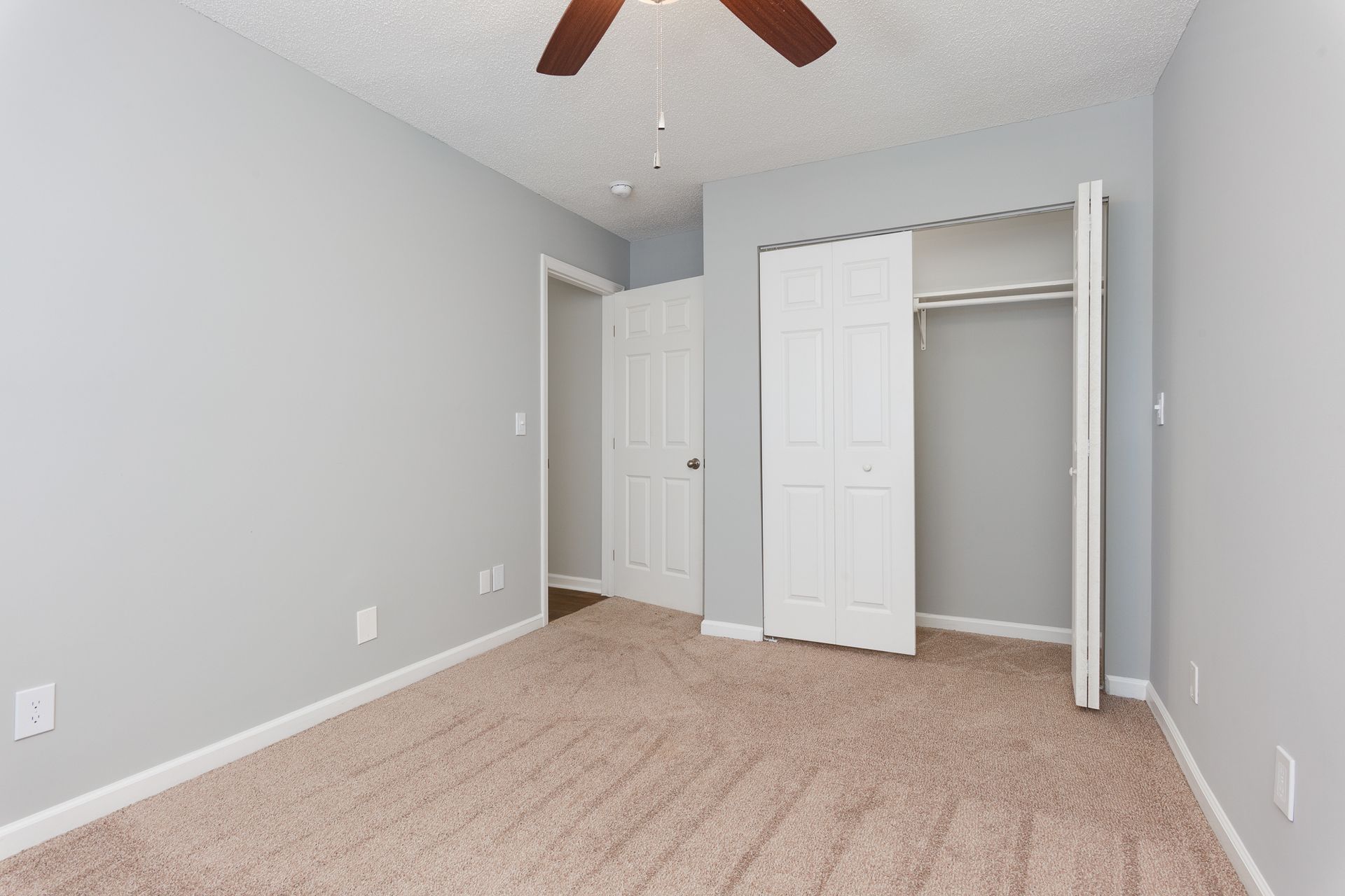 An empty room with light gray walls, beige carpeting, a ceiling fan, a doorway, and an open white closet.