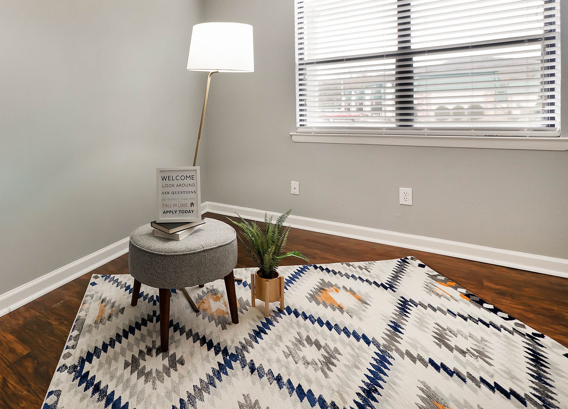 A gray stool with a small sign sits on a patterned rug next to a floor lamp and potted plant in a room with a window.