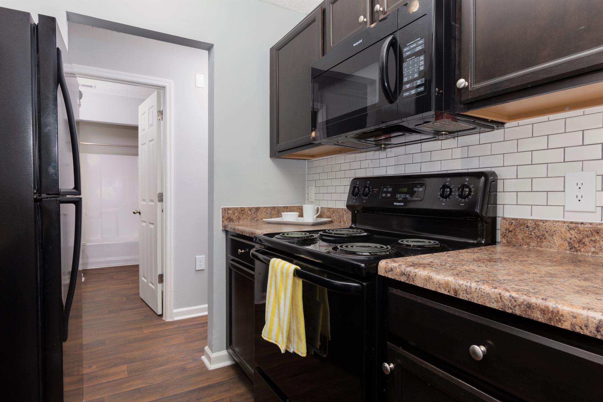 A kitchen featuring dark cabinets, a black stove and microwave, brown speckled countertops, and a white subway tile backsplash.