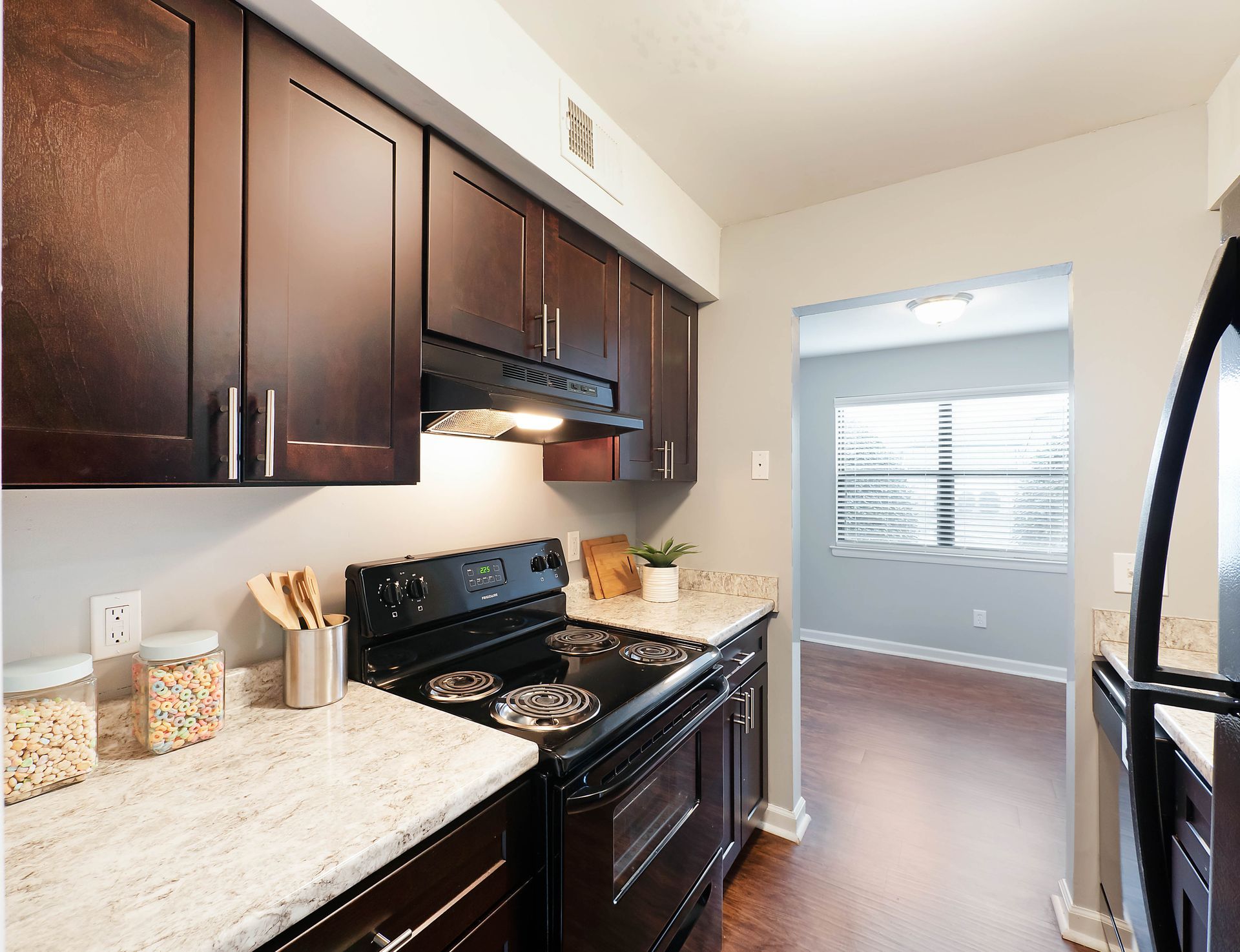 A kitchen with dark wood cabinets, light granite countertops, a black stove, and a doorway leading to a dining area.