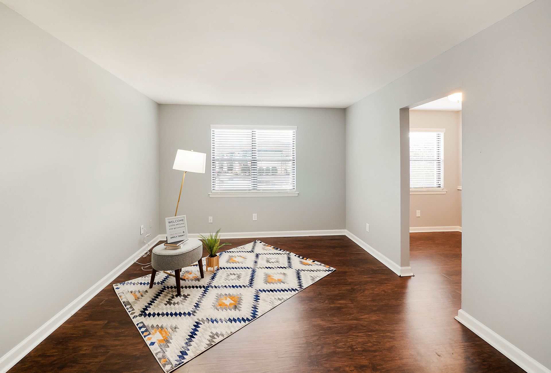 A sparsely furnished room with light gray walls, dark wood floors, a patterned rug, a small stool, and a floor lamp.