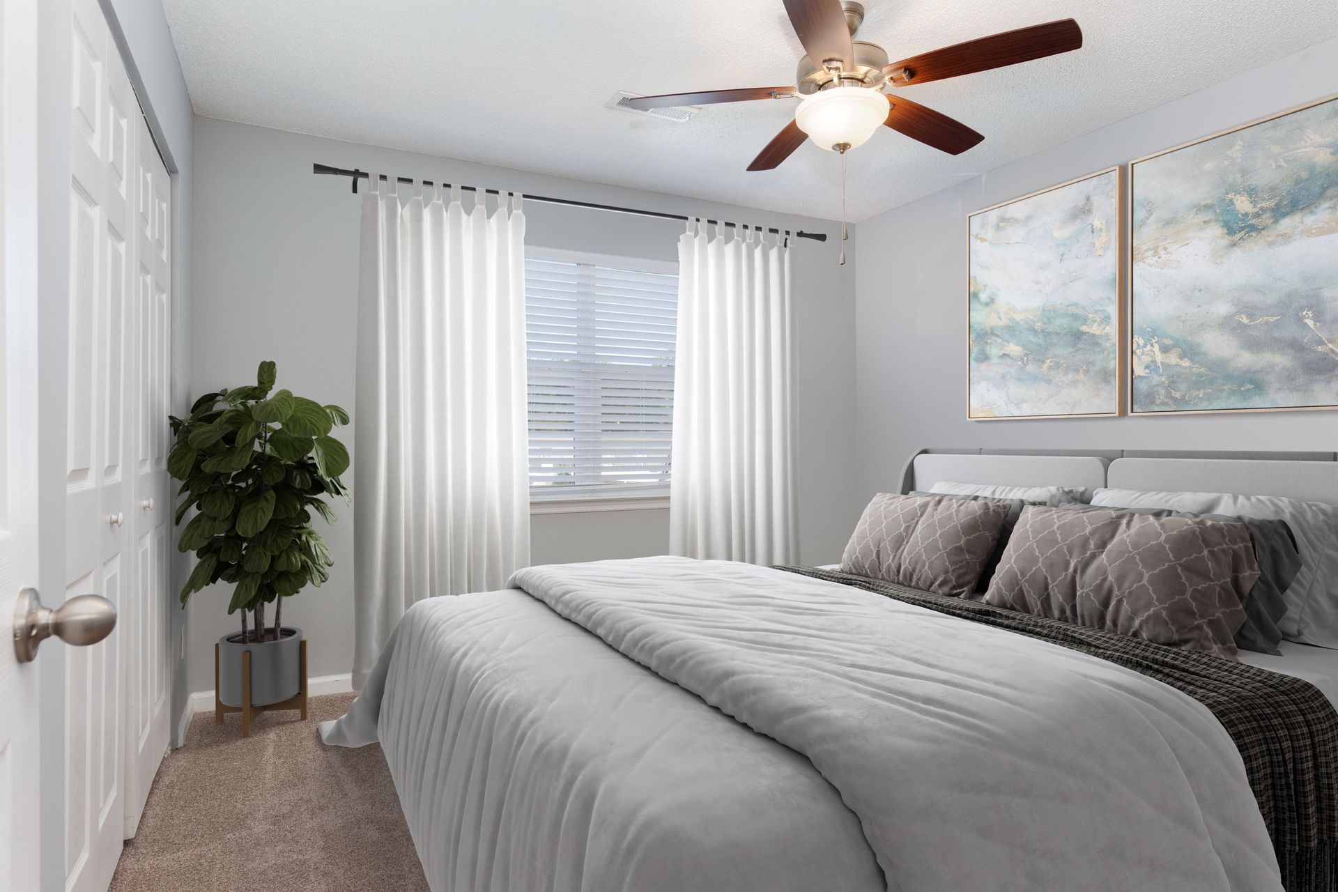 A modern bedroom with a bed featuring a gray comforter, white curtains over a window, a houseplant, and ceiling fan.