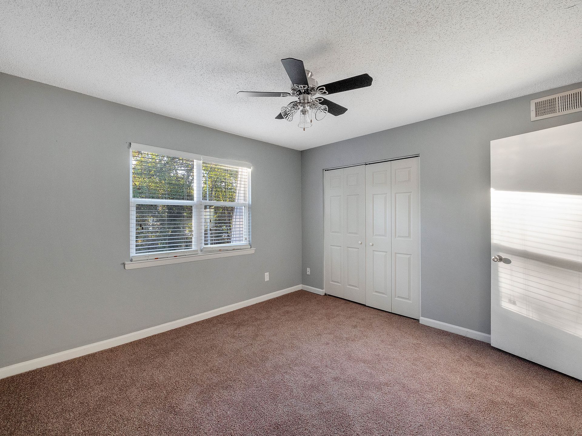 An empty bedroom with light gray walls, brown carpet, a ceiling fan, a window with white blinds, and white closet doors.