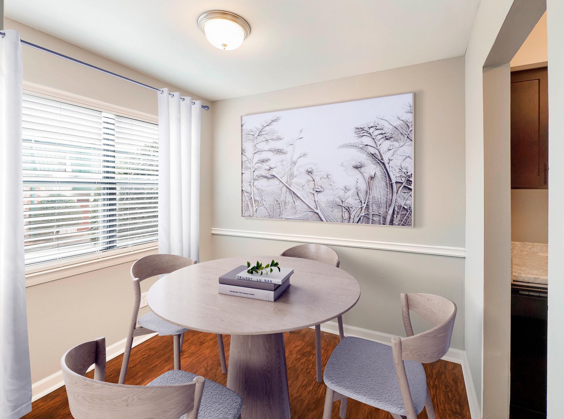 A round dining table with four grey chairs, set in a room with light walls, wood floors, a window, and wall art.
