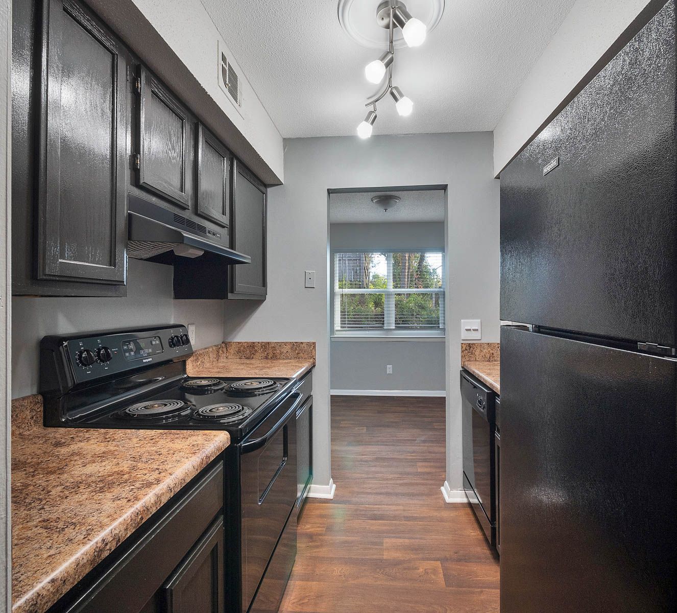 A narrow kitchen with dark cabinets, a black stove, a black refrigerator, brown countertops, and a view into a room.