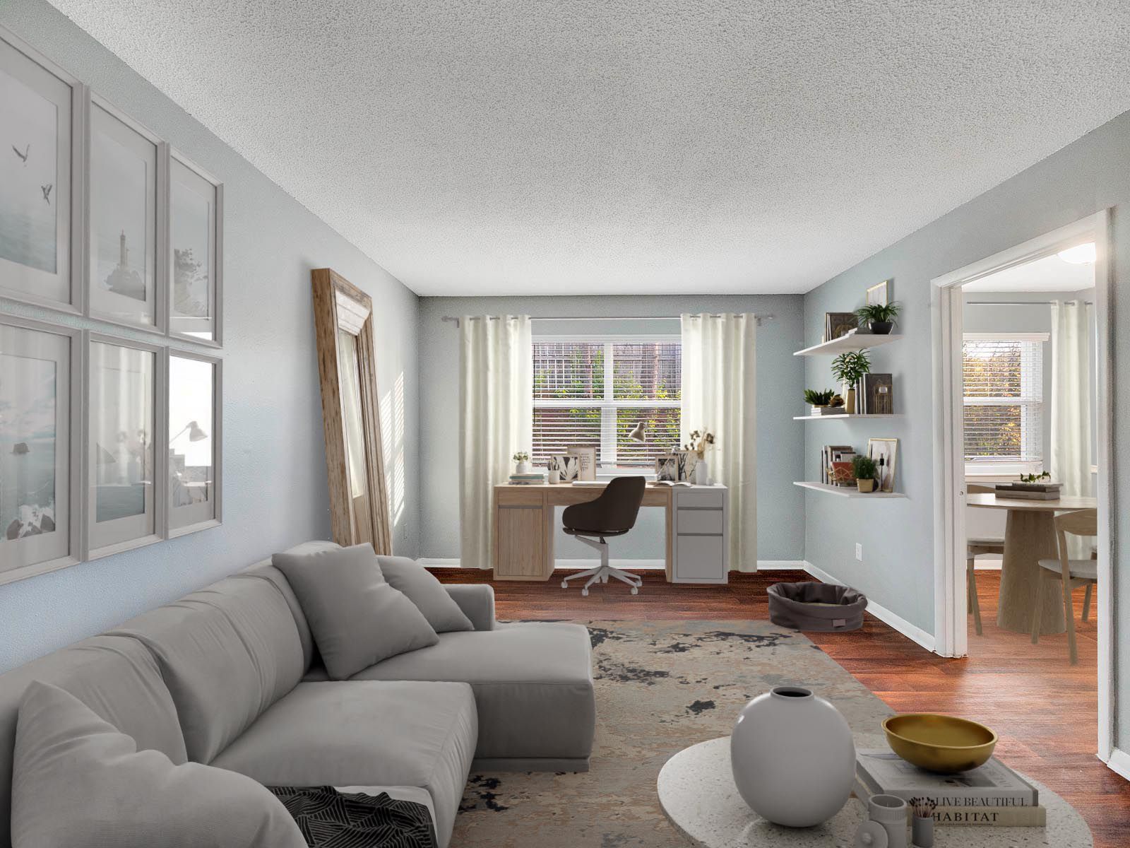 A modern living room with a grey sectional sofa, an office desk by the window, wall shelves, and a view into a dining area.