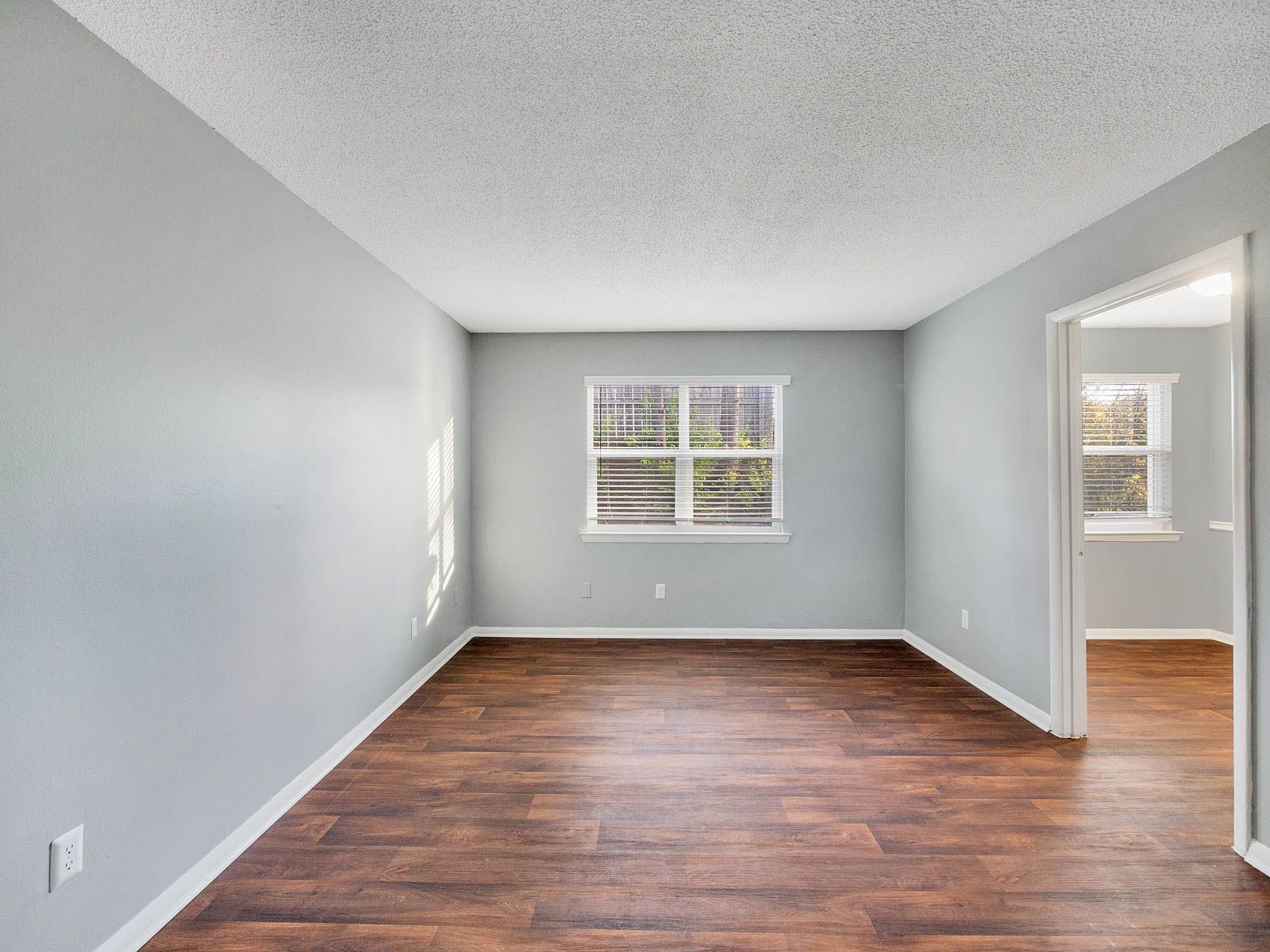 An empty room with light gray walls, dark wood-look floors, a textured white ceiling, and a window viewing greenery.