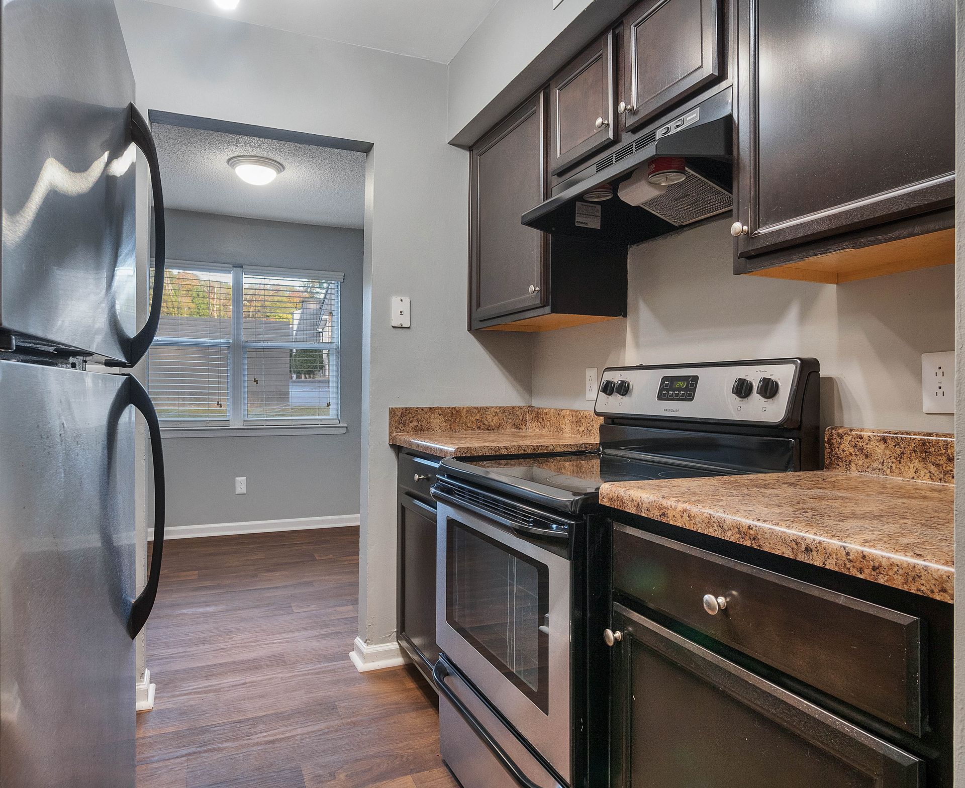A kitchen featuring a stainless steel refrigerator, dark wood cabinets, a tan granite countertop, and a stainless oven.