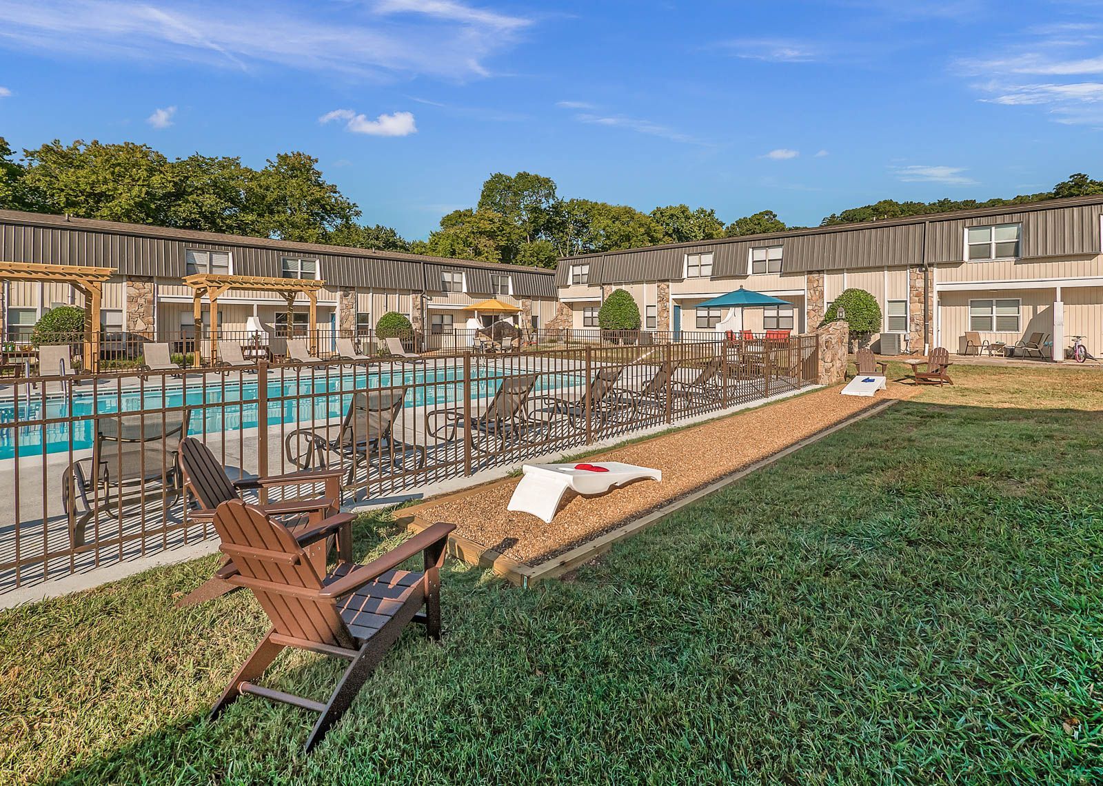 A swimming pool with lounge chairs and wooden pergolas, bordered by a green lawn and multi-story apartment buildings.