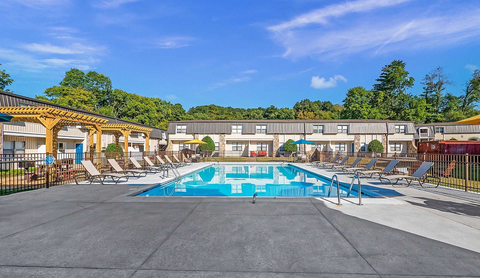 A clear, rectangular swimming pool surrounded by a concrete deck and a wooden pergola under a bright blue sky.