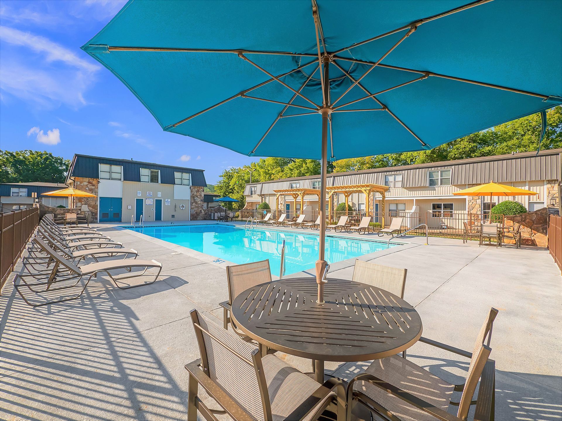 An outdoor swimming pool surrounded by lounge chairs, patio tables with blue and yellow umbrellas, and apartment units.