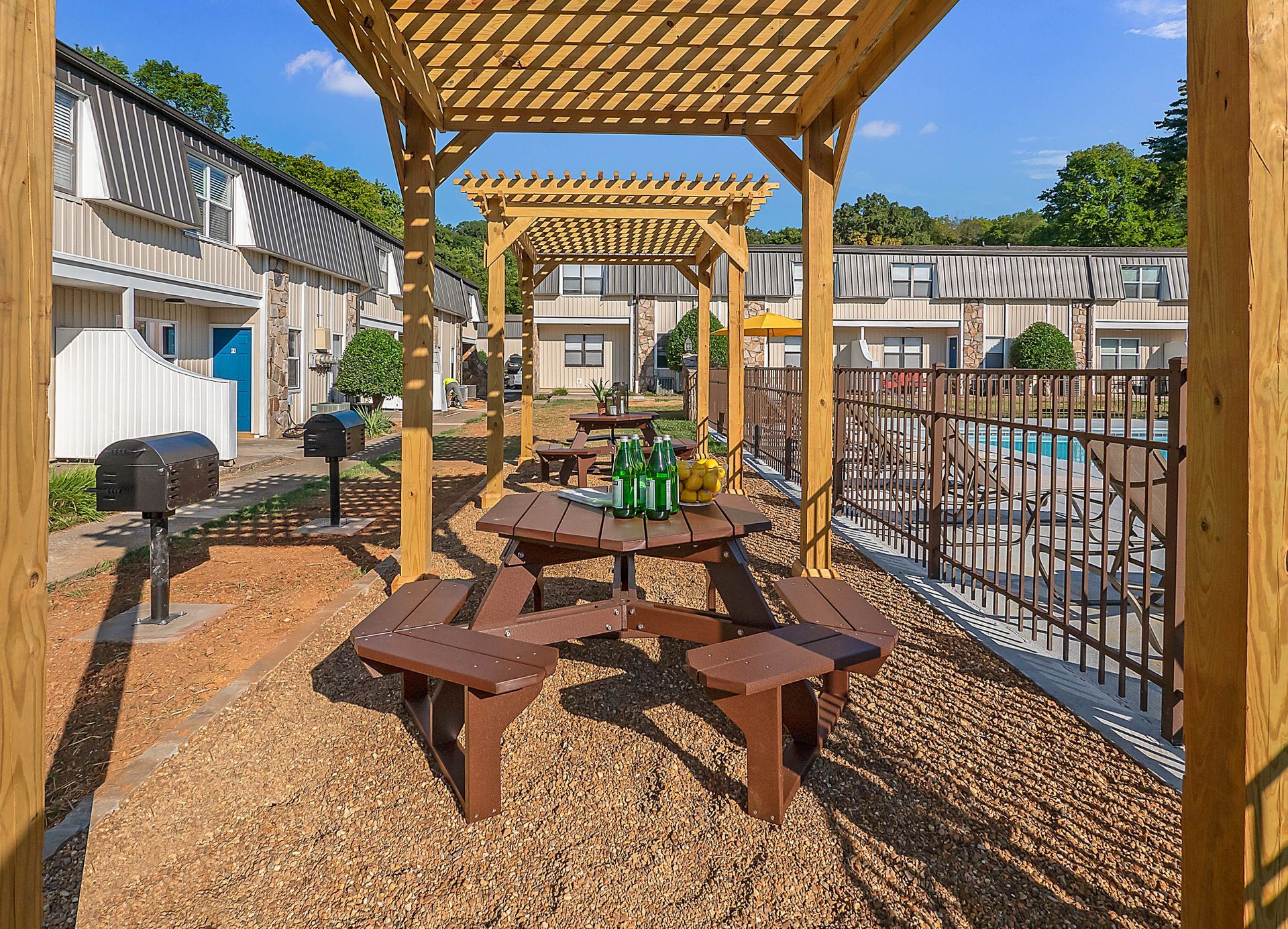 Outdoor communal area with wooden picnic tables under pergolas, surrounded by brown mulch, barbecue grills, and apartments.