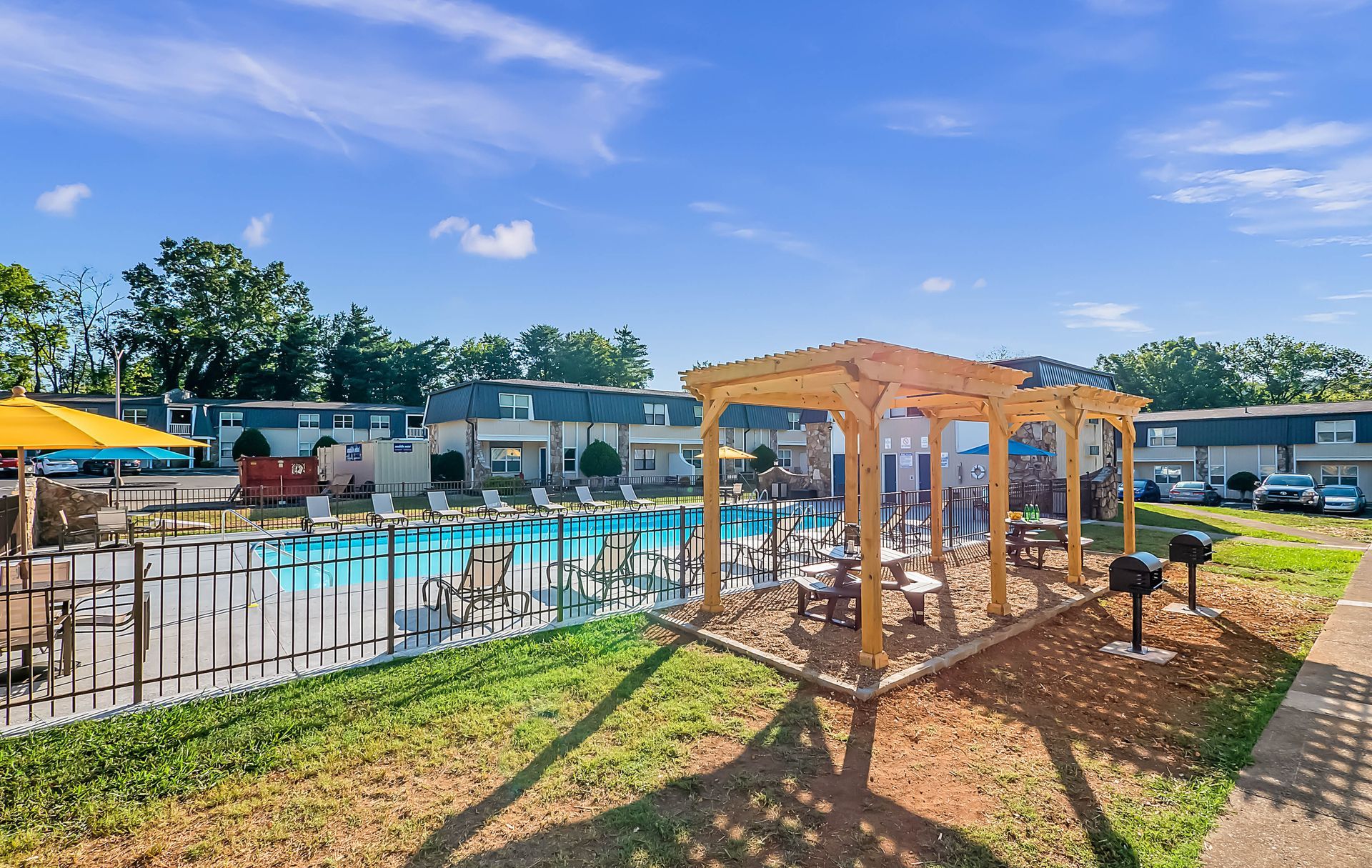 Outdoor swimming pool area with a wooden pergola, lounge chairs, and grills under a bright blue sky.