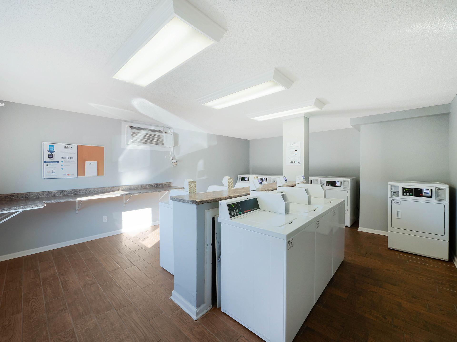 A clean, modern laundry room with white machines, light gray walls, and wood-patterned floors under bright ceiling lights.